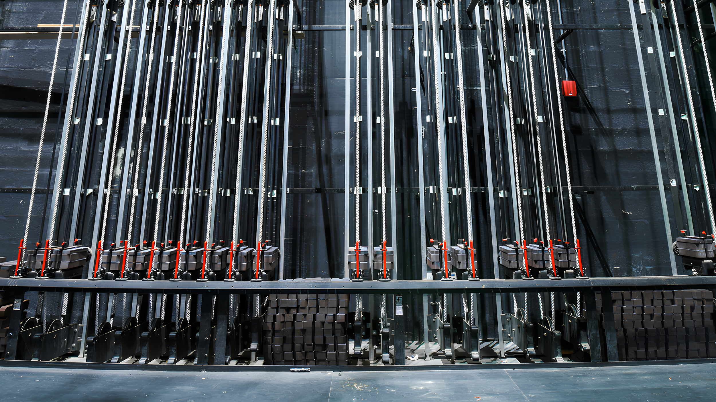 Multiple metal pipes or rods secured in position with clamps and red levers, arranged in rows, mounted on a black surface, with a black wall in the background.