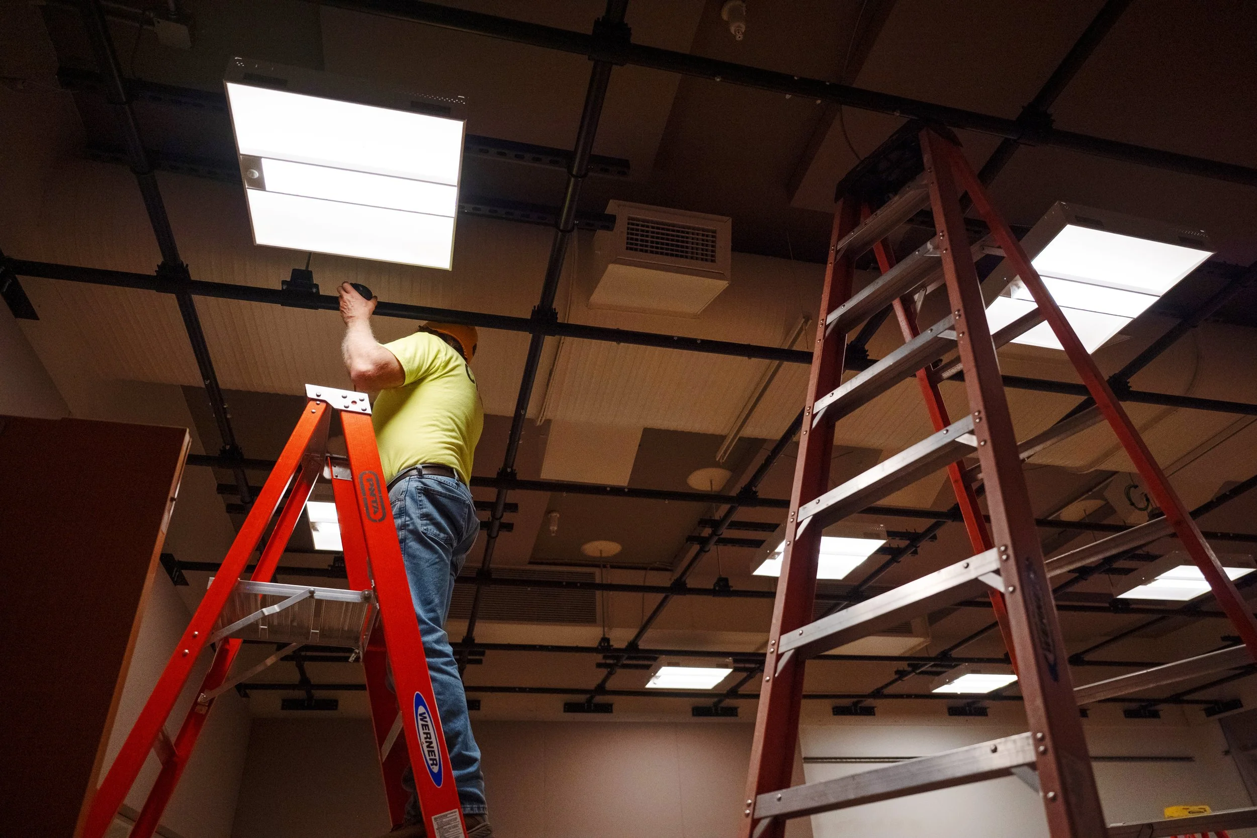 A man standing on a red stepladder installing ceiling panels in a room with a drop ceiling grid, with another stepladder nearby. The room has a brown ceiling, some ceiling lights, an air vent, and dark walls.