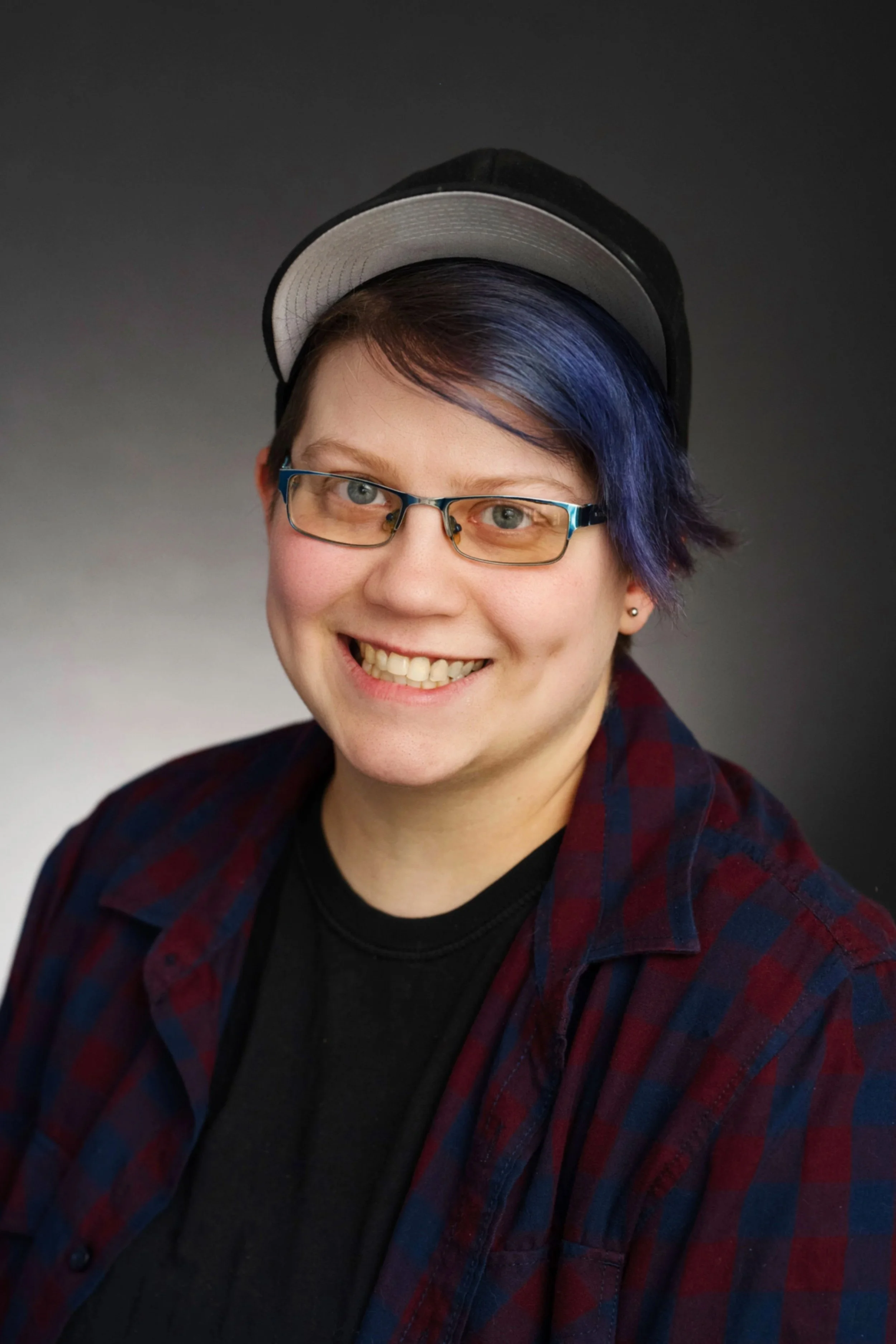 A smiling person with short brown hair, glasses, and blue highlights wearing a black shirt and a red and blue checkered flannel shirt, with a dark gradient background.