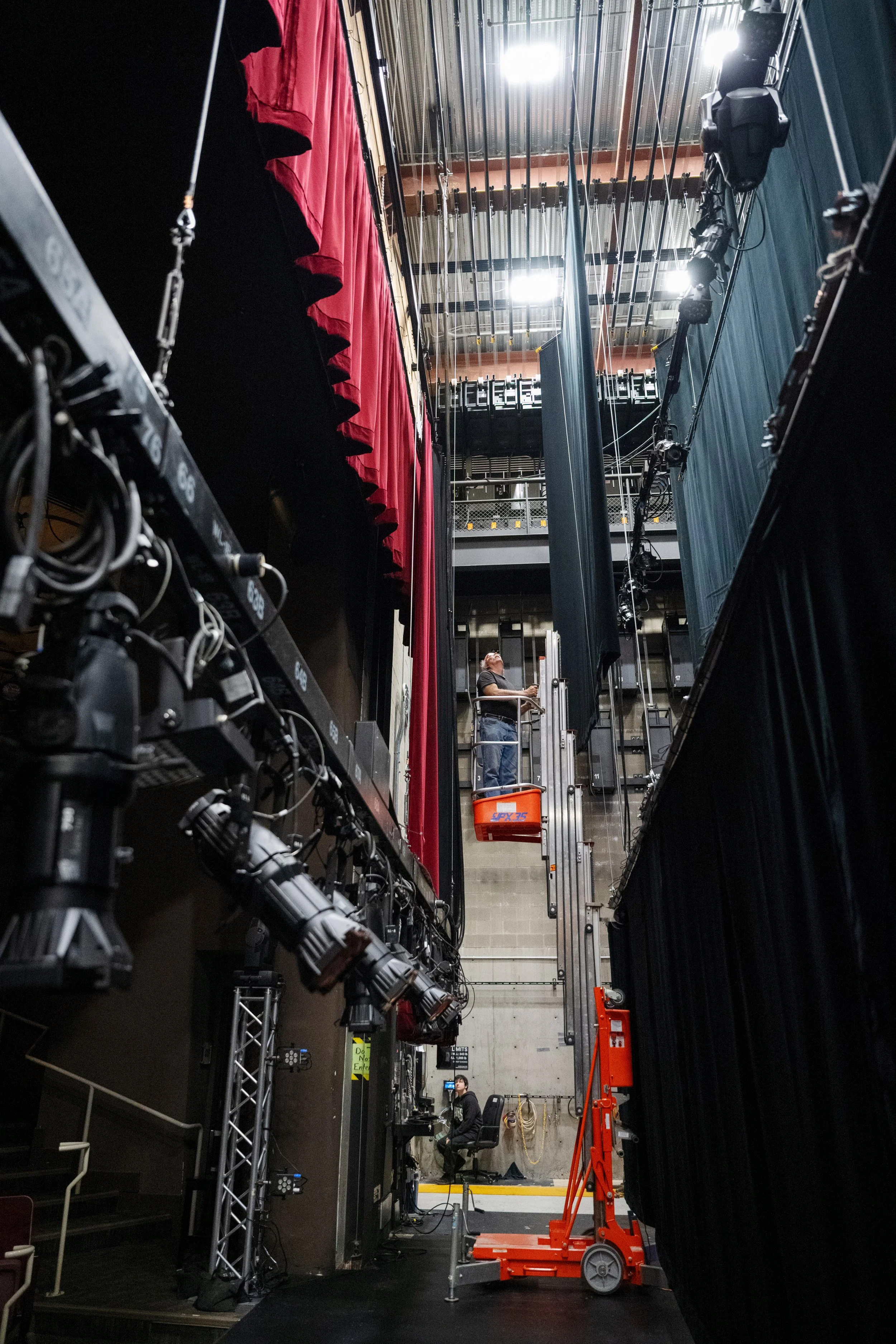 View of theater stage from backstage, showing stage lights, curtains, and stage equipment such as a lift with a person operating it.