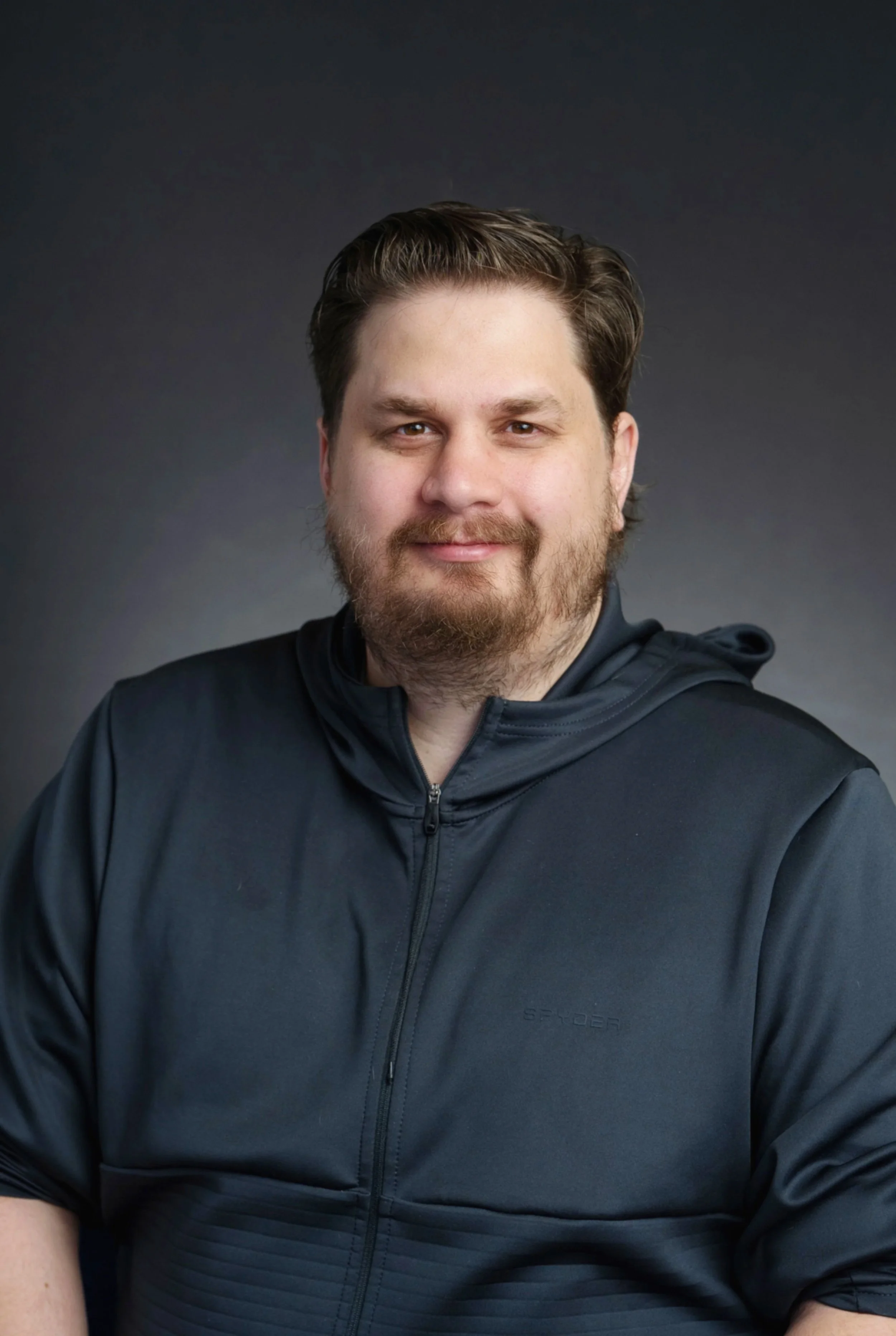 A man with short brown hair, a beard, and light skin, wearing a black zip-up jacket, smiling slightly, against a dark gray gradient background.