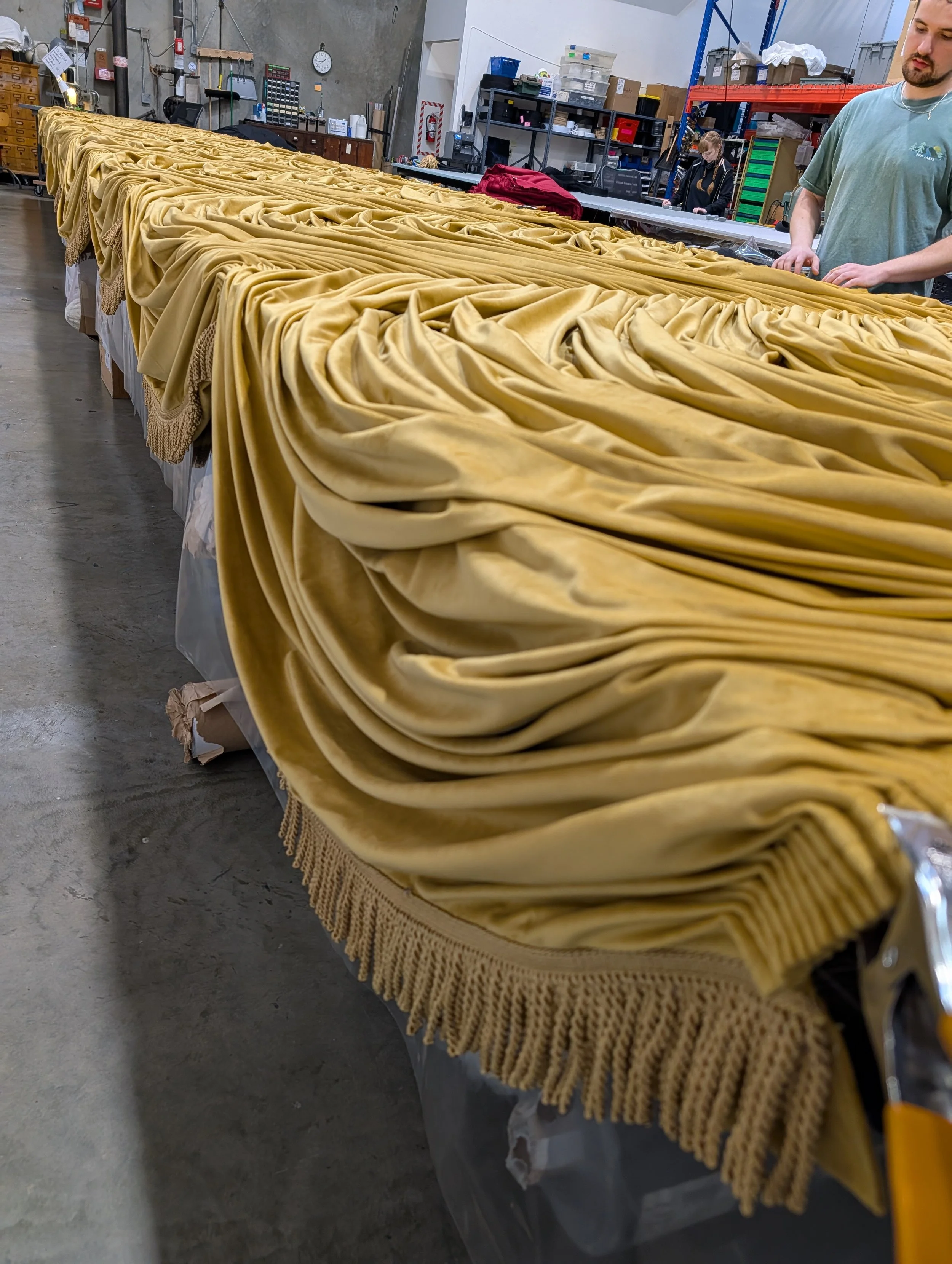 A long table covered with numerous yellow velvet curtains with gold fringe, in an industrial workshop setting.