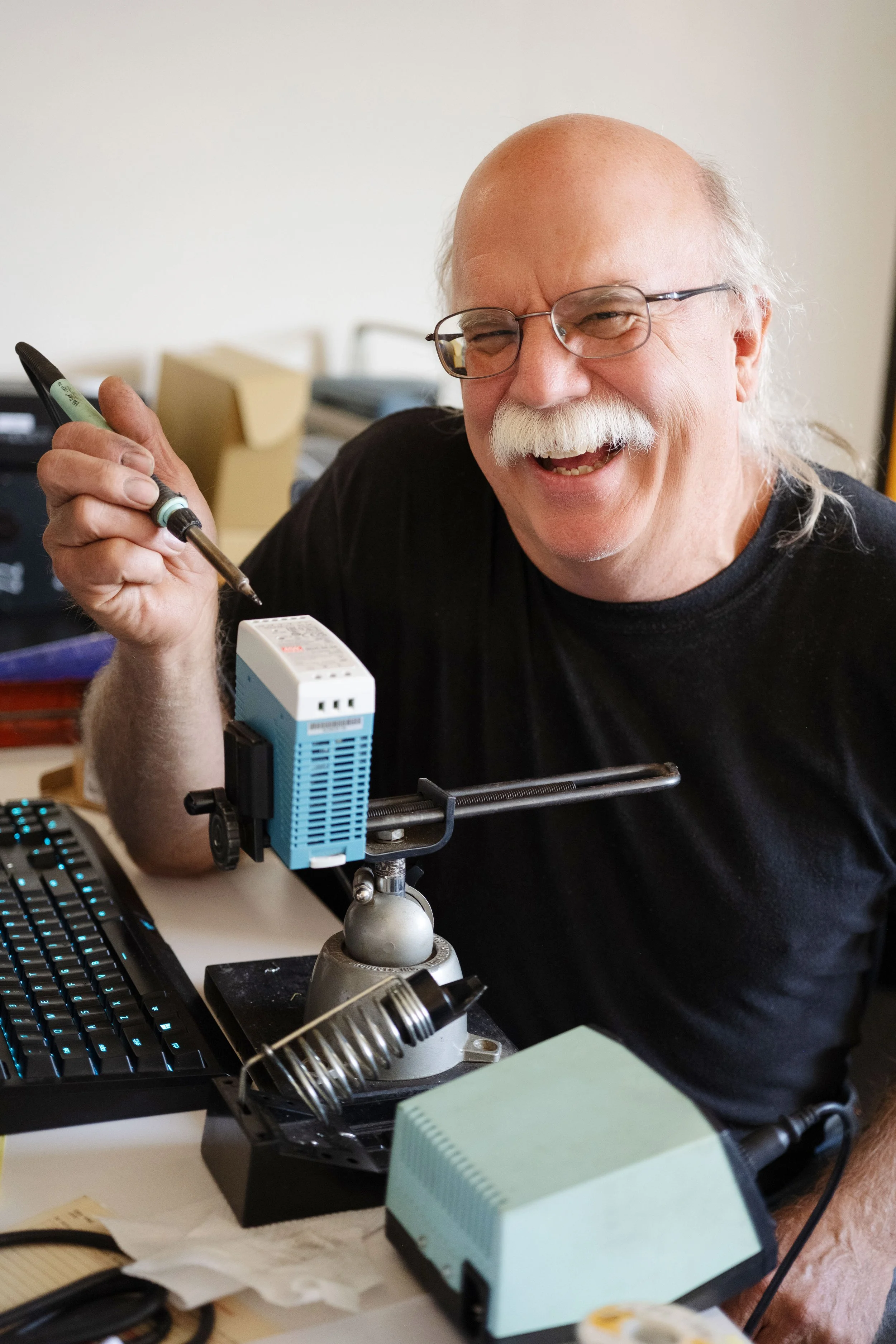 An older man with glasses and a white mustache smiling while working at a desk with various electronic and mechanical components, including a soldering iron, a small blue electronic device, and a computer keyboard.