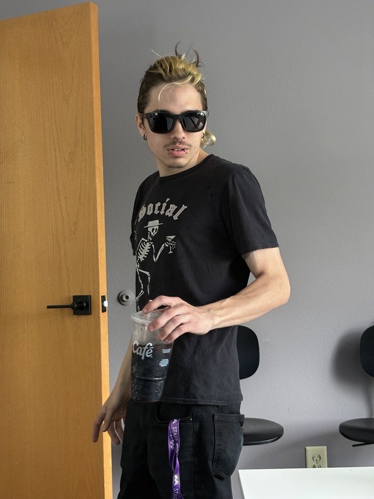 A young man with blond and dark hair, wearing large black sunglasses, a black T-shirt with a skeleton graphic and the word 'social,' and black pants with a purple keychain, standing in a room with a gray wall, black chairs, and a wooden door, holding a cup of dark soda.