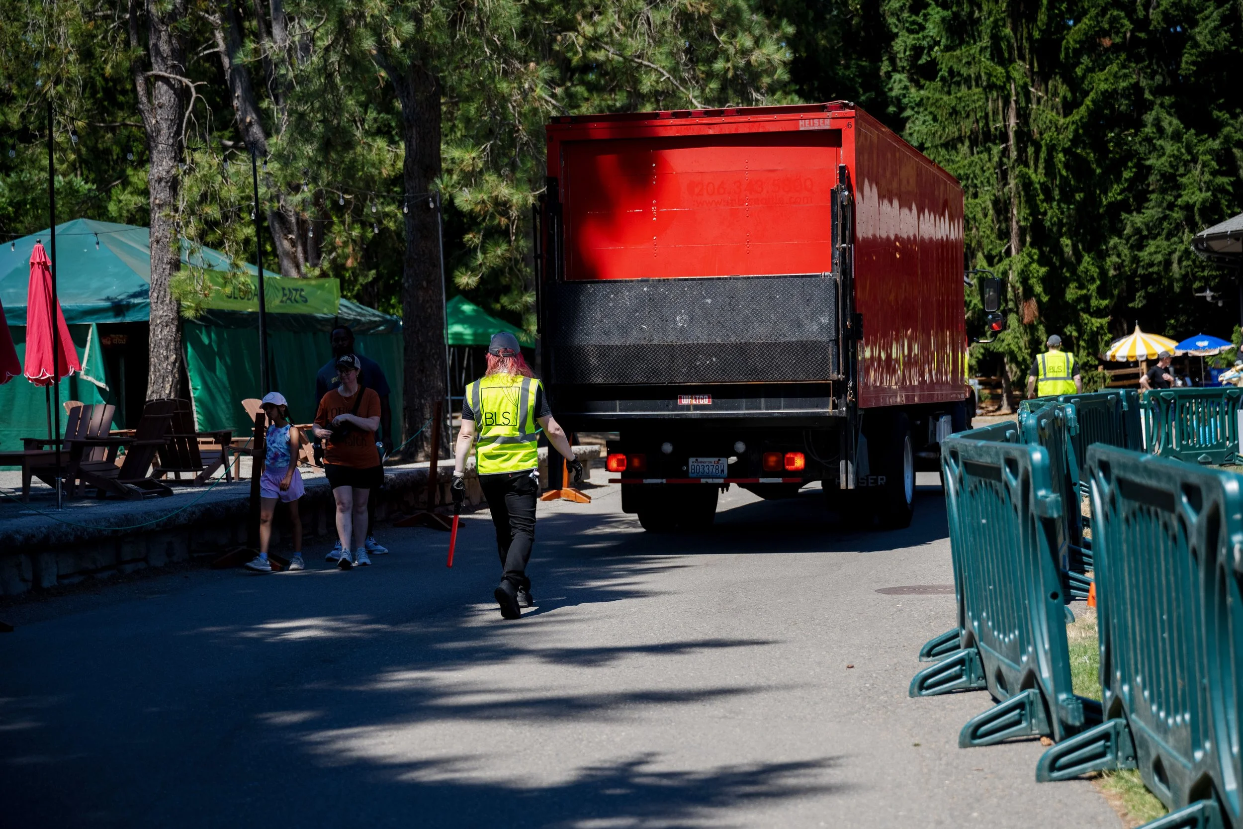 A red delivery truck parked on a street during daytime, with people walking nearby and green trees in the background. Some people are wearing safety vests, and tents or booths are set up on the sidewalk.