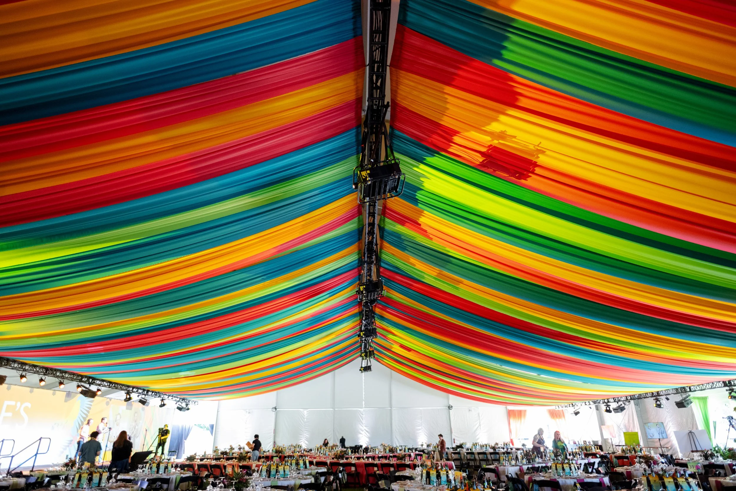 Large indoor event space with a colorful fabric canopy ceiling in shades of red, orange, yellow, green, and blue, mounted on a truss structure. The space has tables and chairs set up for an event, with people walking around and preparing.