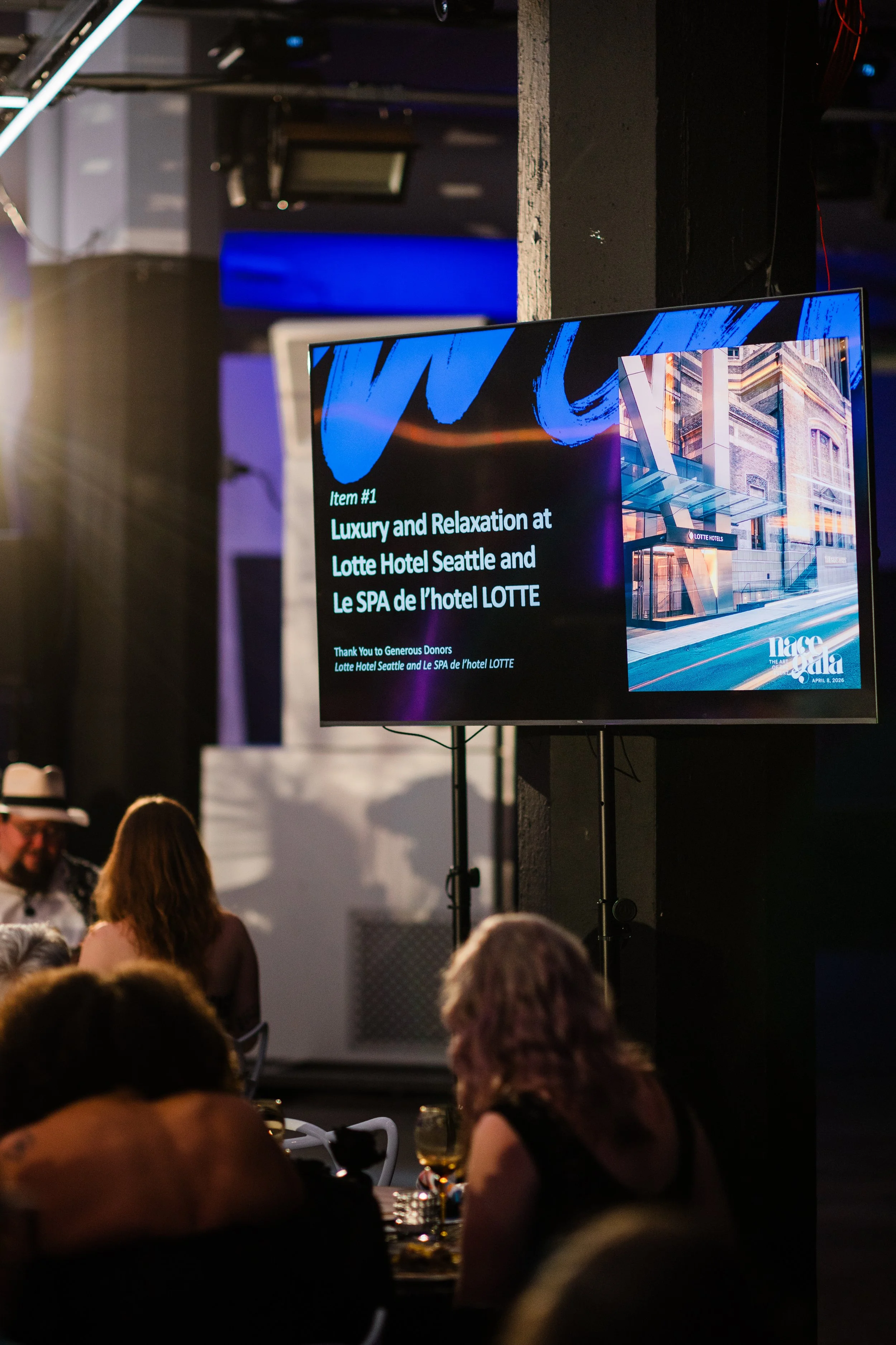 A presentation slide on a large screen in a dimly lit venue showing information about luxury and relaxation at Lotte Hotel Seattle. The slide includes a photo of a modern hotel building and a message thanking donors.