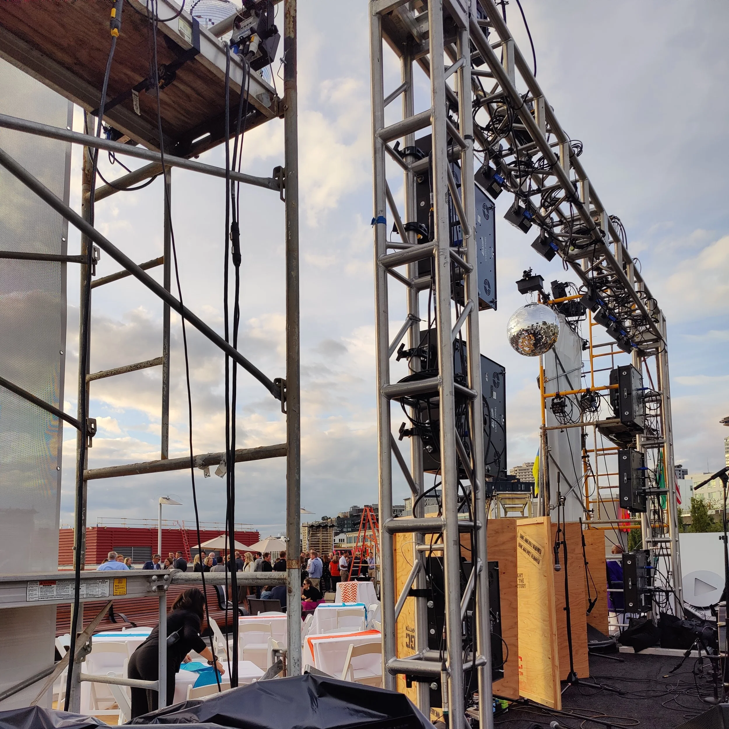 Outdoor event with scaffolding, audio equipment, and a group of people gathered, likely preparing for a performance or ceremony, with buildings and cloudy sky in the background.