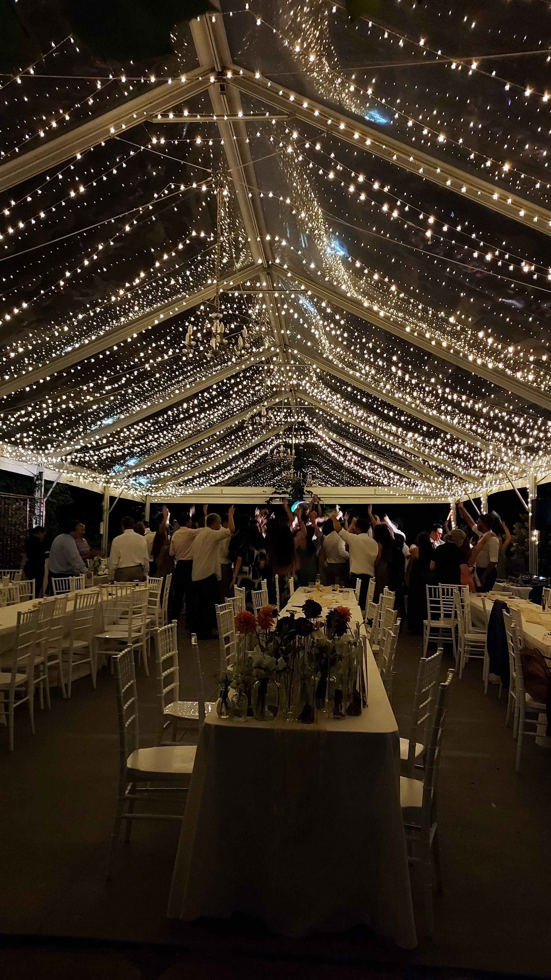 A nighttime outdoor event with a tent decorated with numerous string lights, and guests dancing underneath. The scene includes tables, chairs, and floral arrangements.