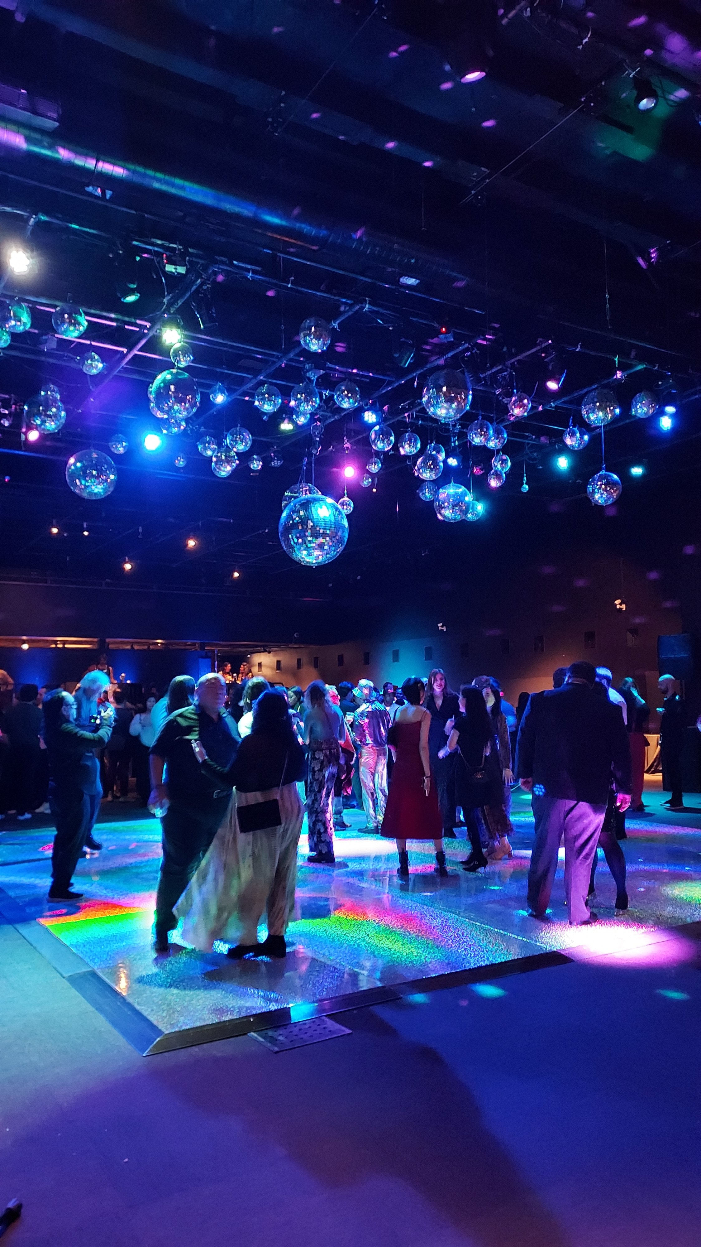 People dancing in a dark, colorful club with disco balls and vibrant lighting.