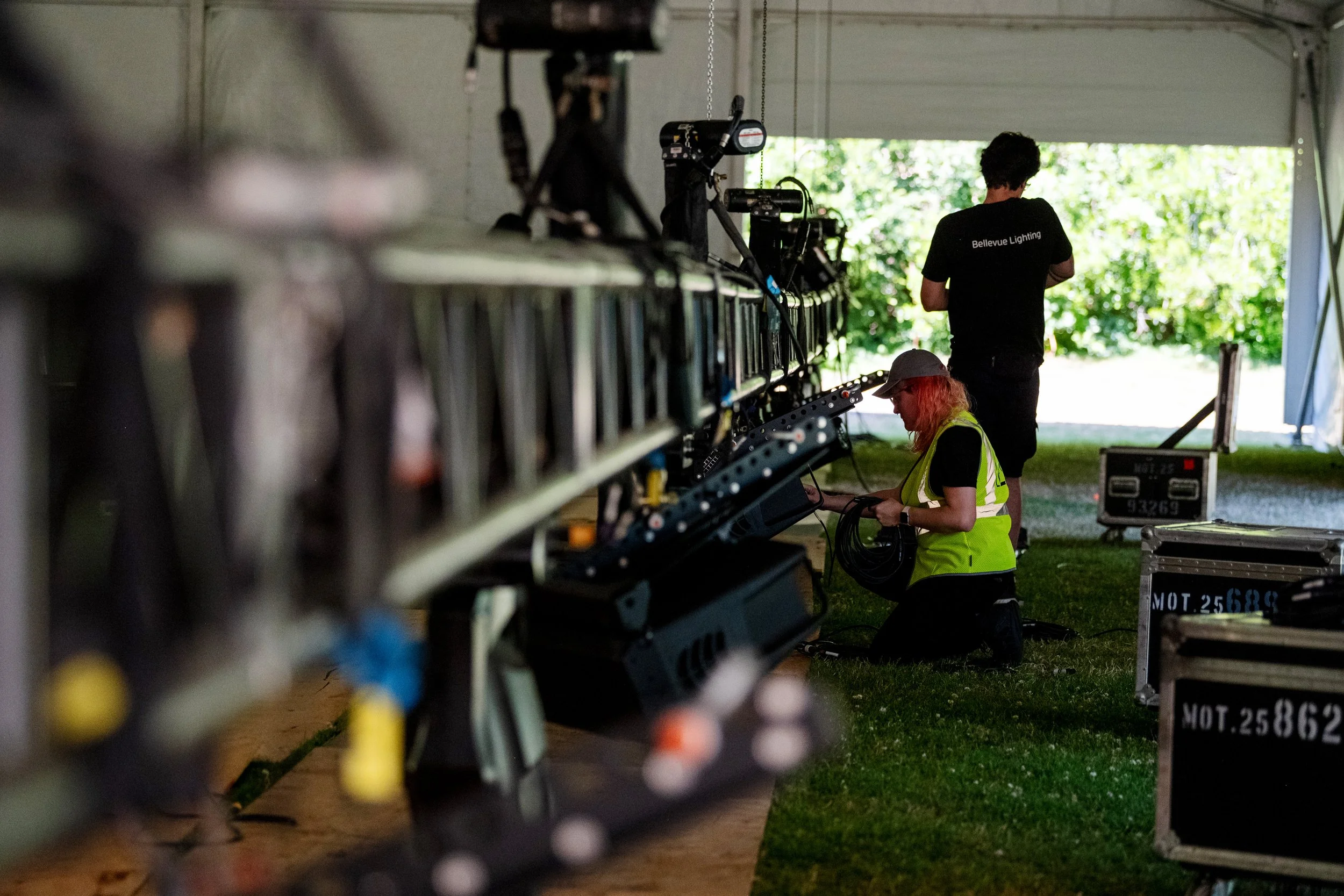 Workers setting up lighting and sound equipment inside a large tent or garage for an event. One worker in a high-visibility vest is kneeling and handling cables, while another stands with his back to the camera, wearing a black t-shirt that reads 'Bellevue Lighting'.