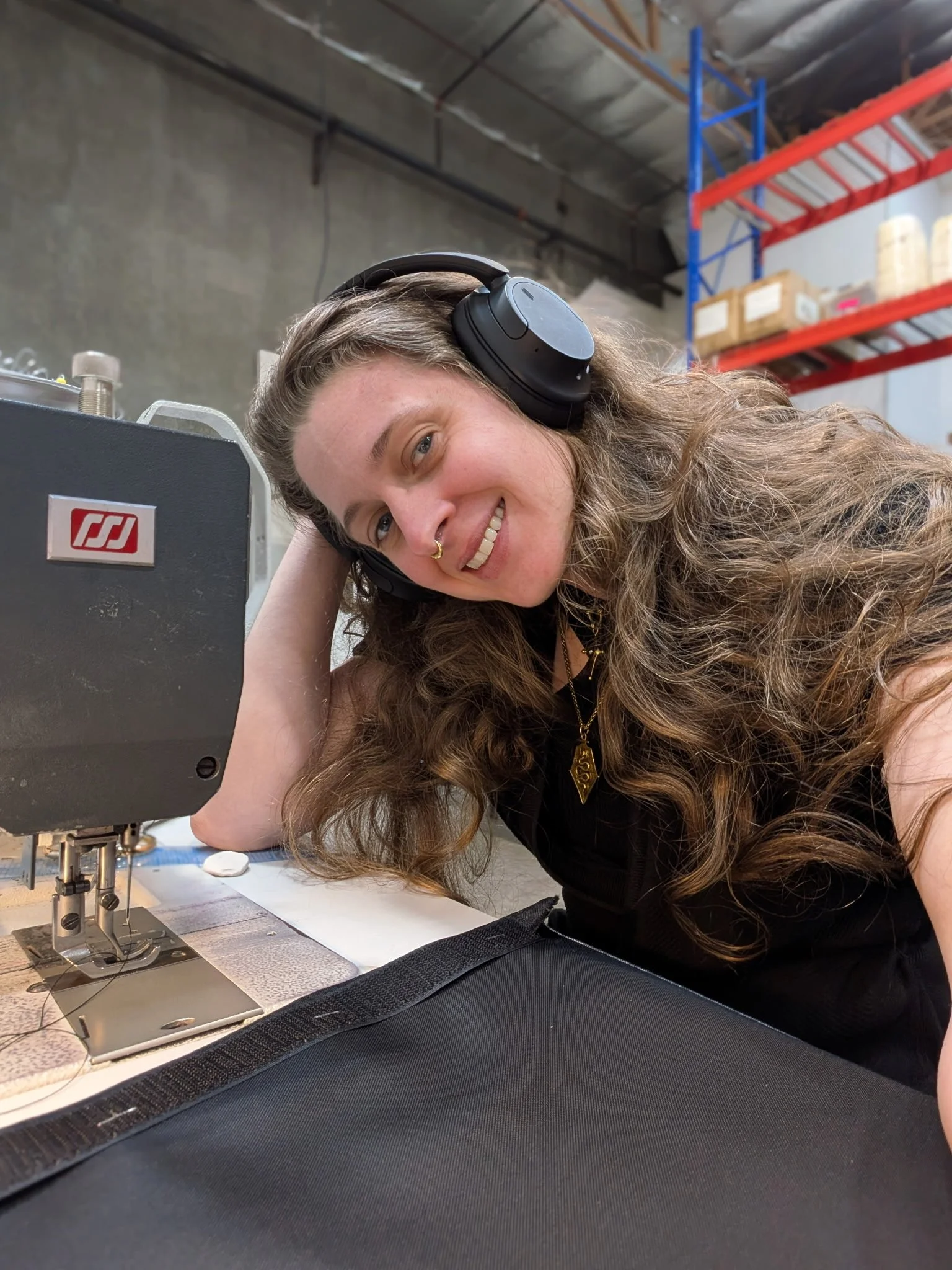 A woman with long curly hair, wearing headphones, smiling, and resting her head on her arm while sitting at a sewing machine in a workshop or industrial setting.