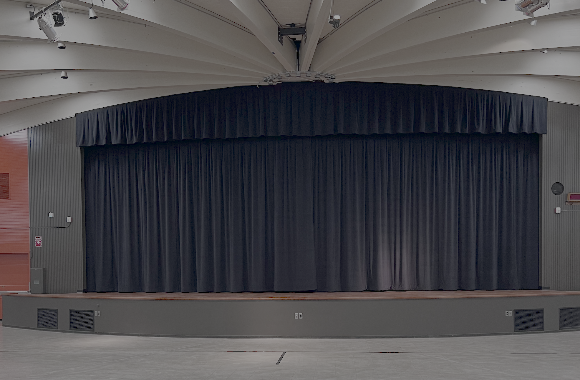 Empty stage with a closed black curtain, set in a modern auditorium with overhead lights and a curved ceiling.