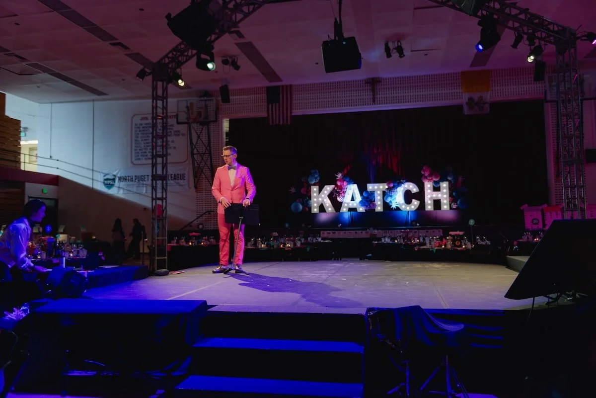Man in pink suit standing on stage with large lit-up letters spelling 'KATCH' behind him at an indoor event venue with decorations, lighting, and tables set up.