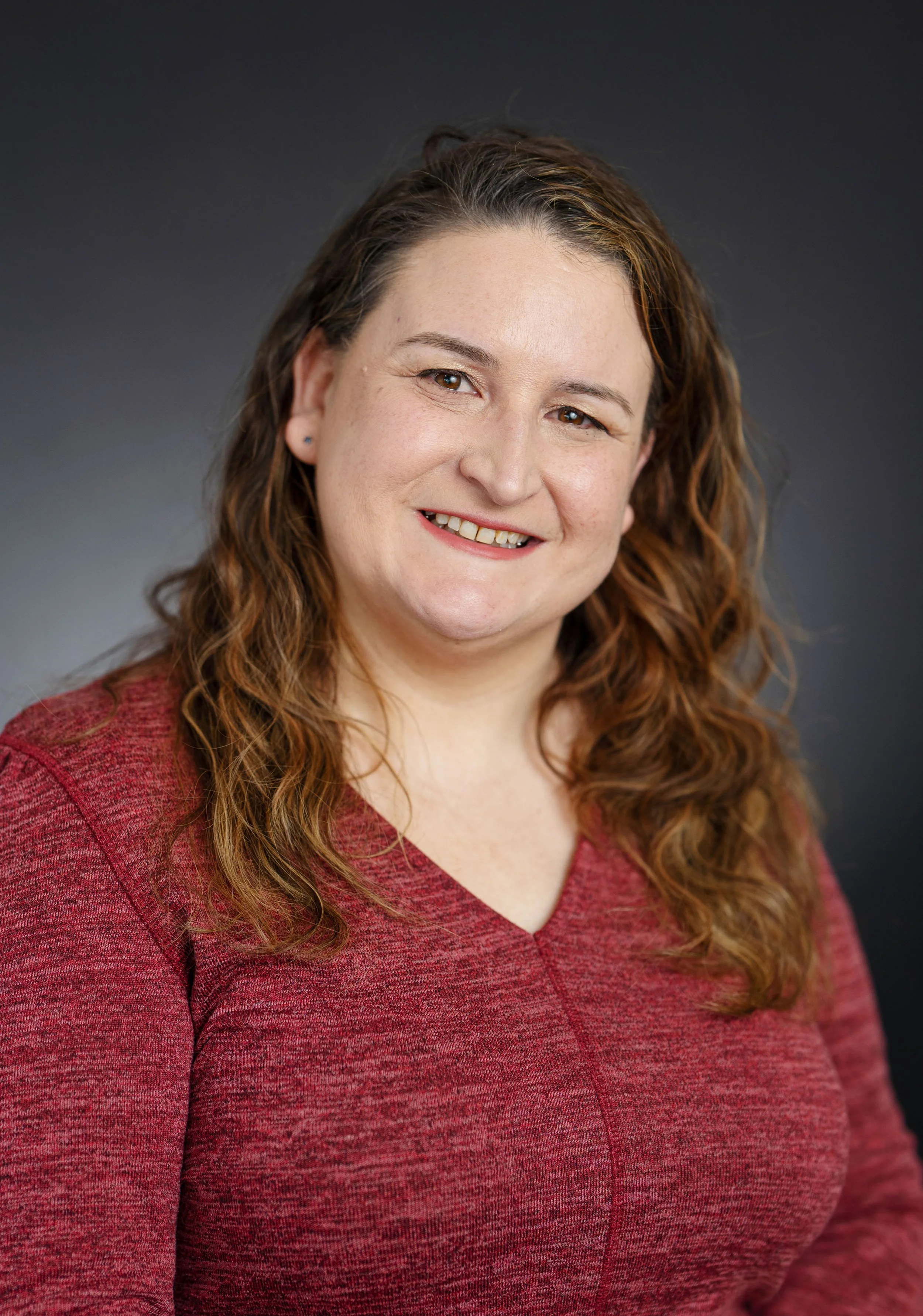 A woman with shoulder-length curly brown hair, wearing a red V-neck sweater, smiling against a dark gray background.