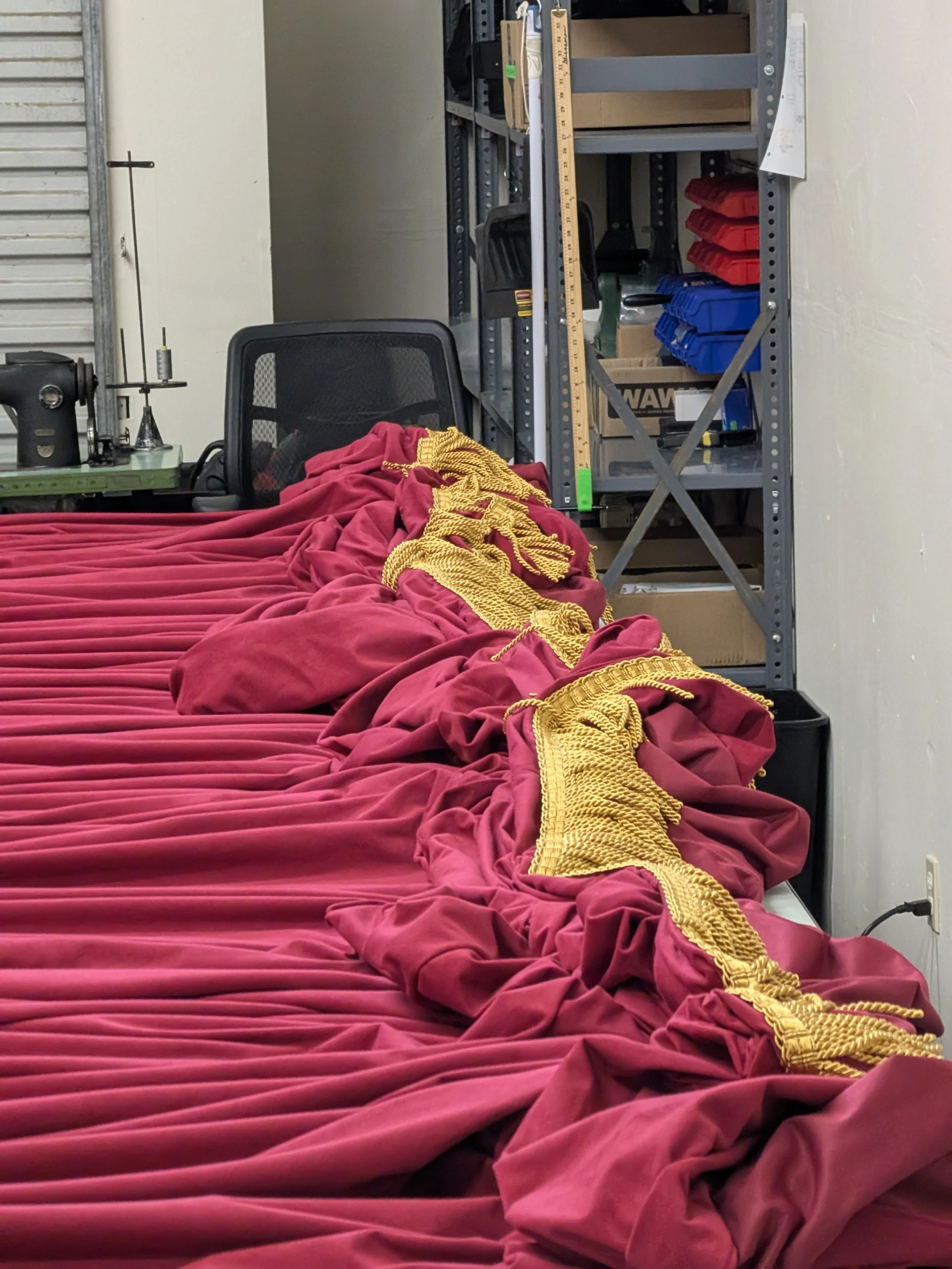 A workbench covered with a red velvet cloth with a gold decorative rope, a black office chair, a metal shelving unit with boxes and tools, and a yellow ruler hanging on the shelf.