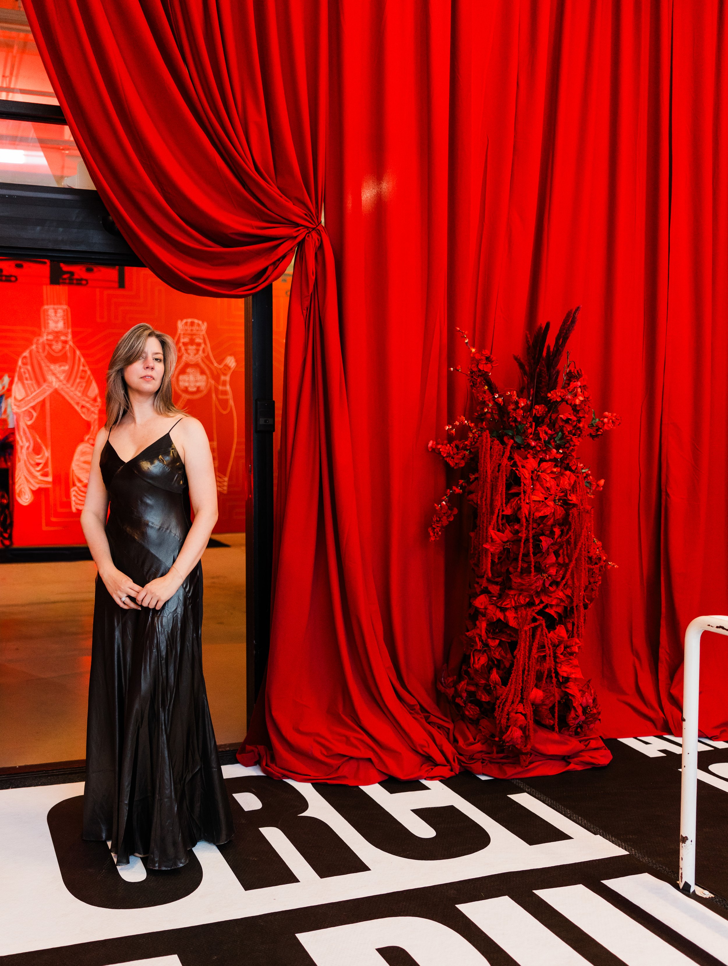 A woman wearing a black satin maxi dress standing in front of a red curtain with floral arrangement to her right.