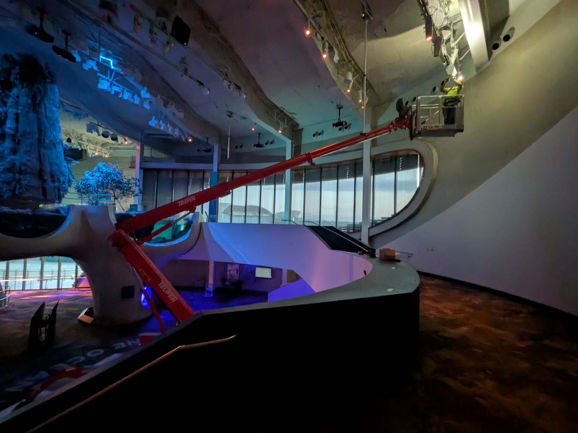 An indoor scene featuring a large white futuristic sculpture of a whale, with a red crane lift working on or near it. There are large windows in the background, some illuminated decorations, and a high ceiling with various lighting fixtures.