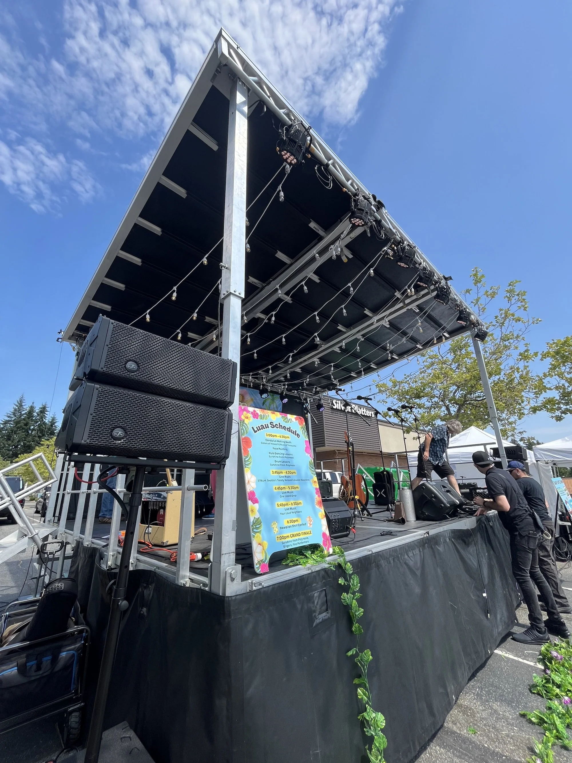 Stage set up outdoors with a black background, large speakers, and a colorful schedule poster, with some people adjusting equipment and a blue sky with clouds overhead.