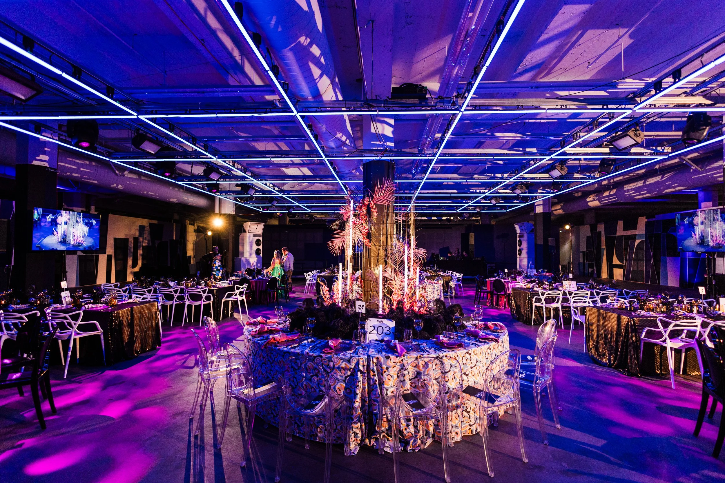 An indoor event space decorated for a party or celebration, with tables set for guests and a large floral centerpiece in the middle. The ceiling has blue neon light fixtures and spotlights, creating a vibrant atmosphere.