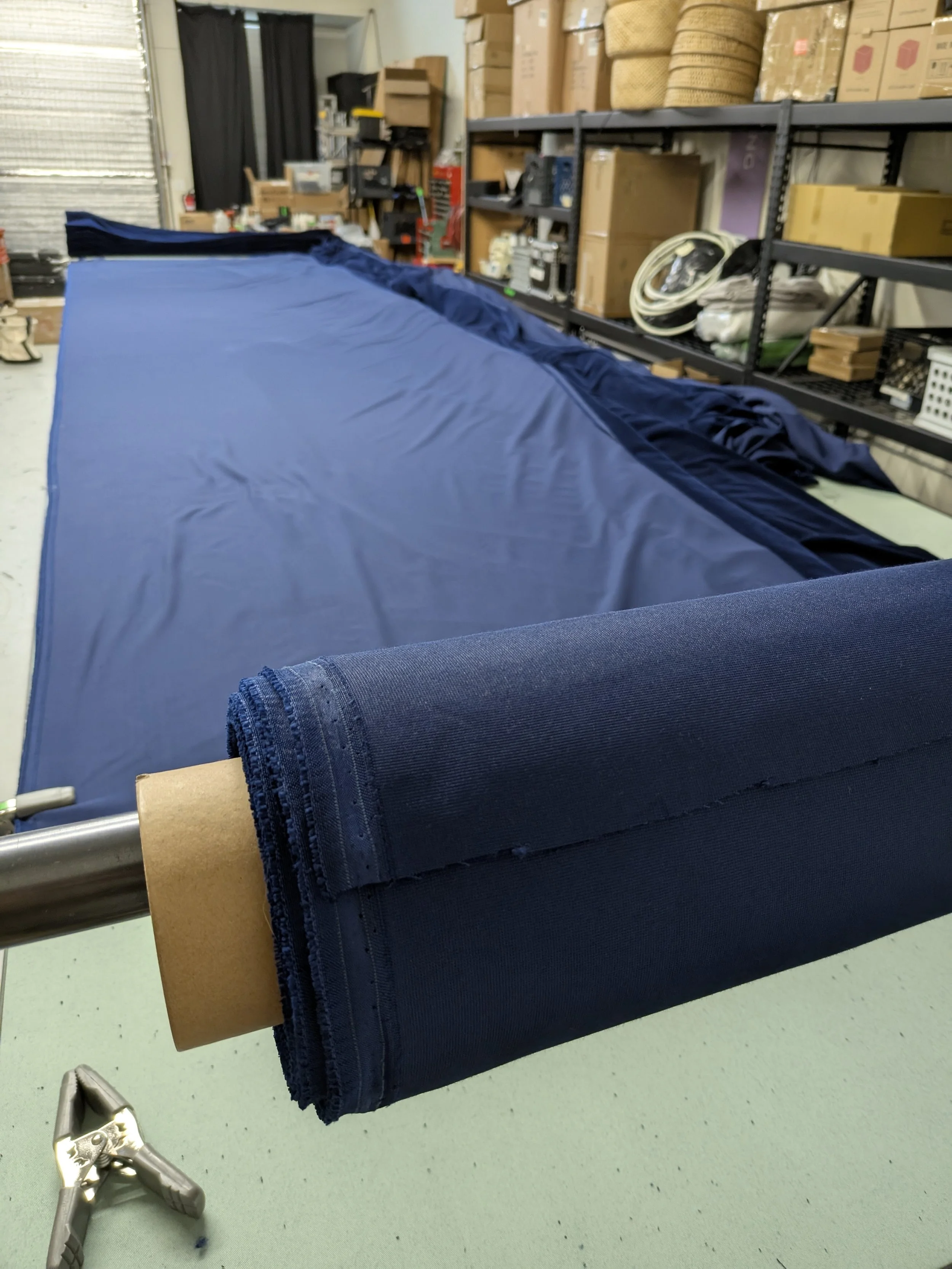 A workshop with a metal table holding a large roll of dark fabric, and a shelving unit with boxes and equipment in the background.