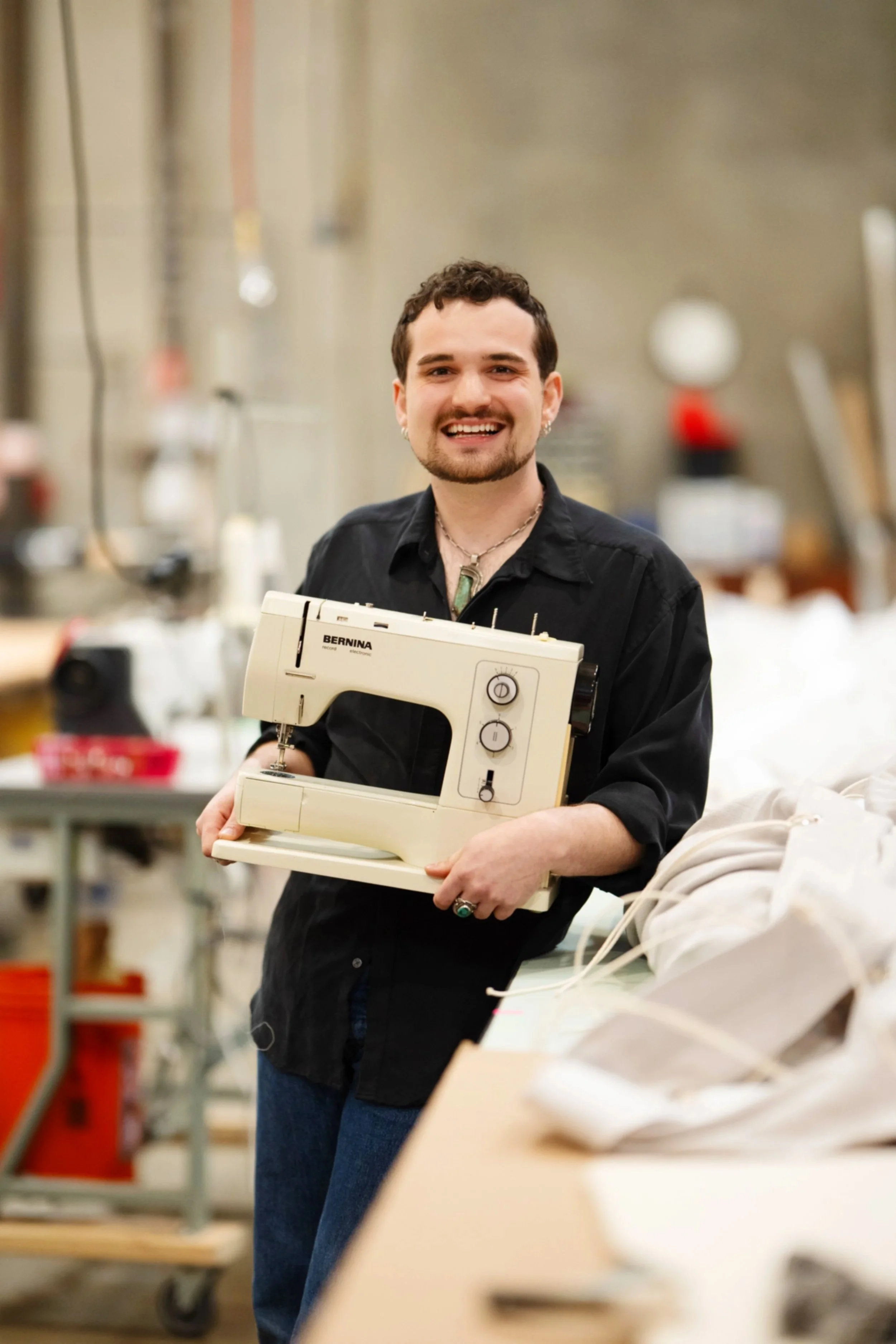 A smiling person holding a sewing machine in an industrial workspace.