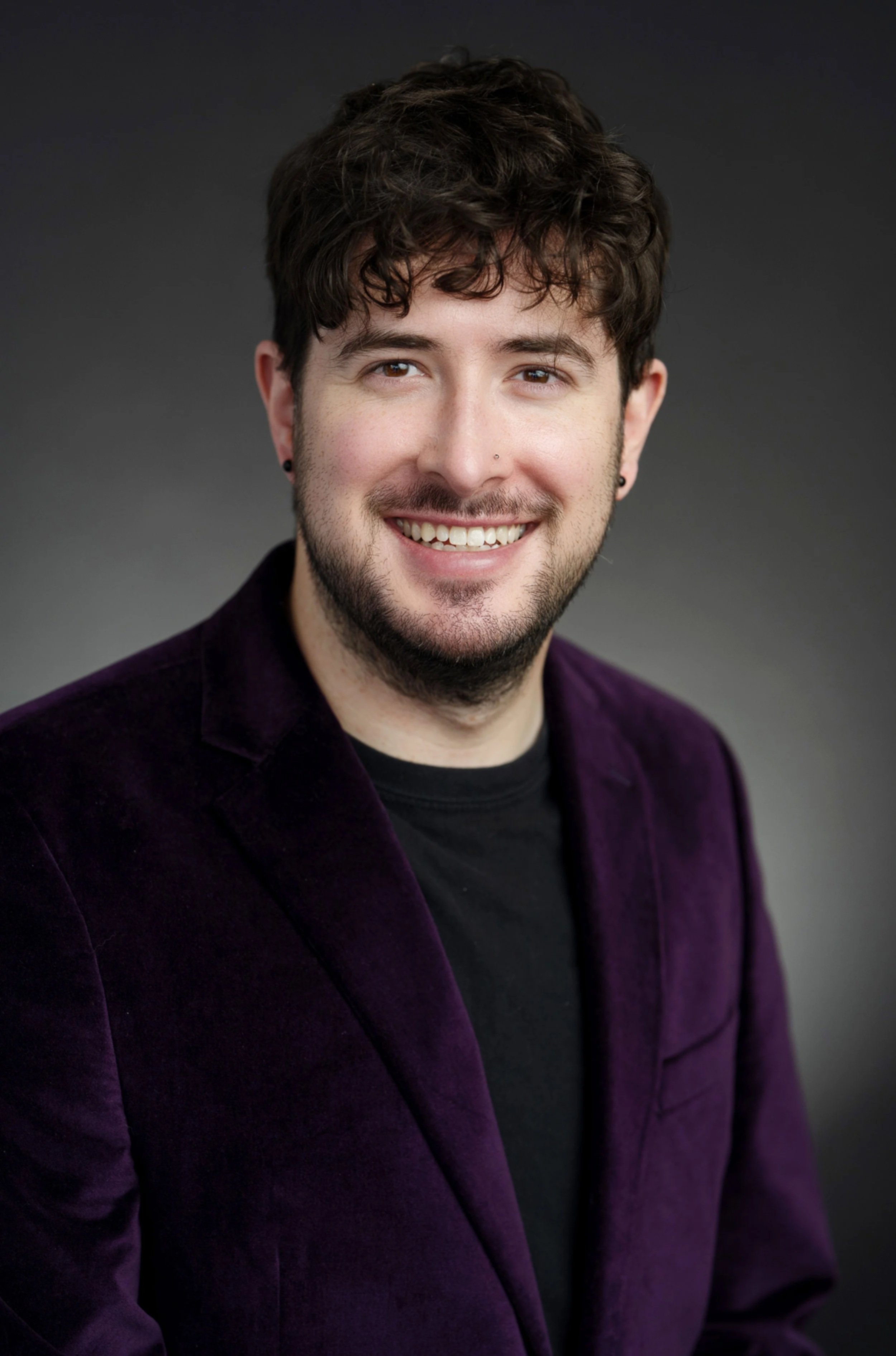 Headshot of a young man with dark wavy hair, brown eyes, a nose piercing, earrings, and a beard, smiling in front of a gray background.