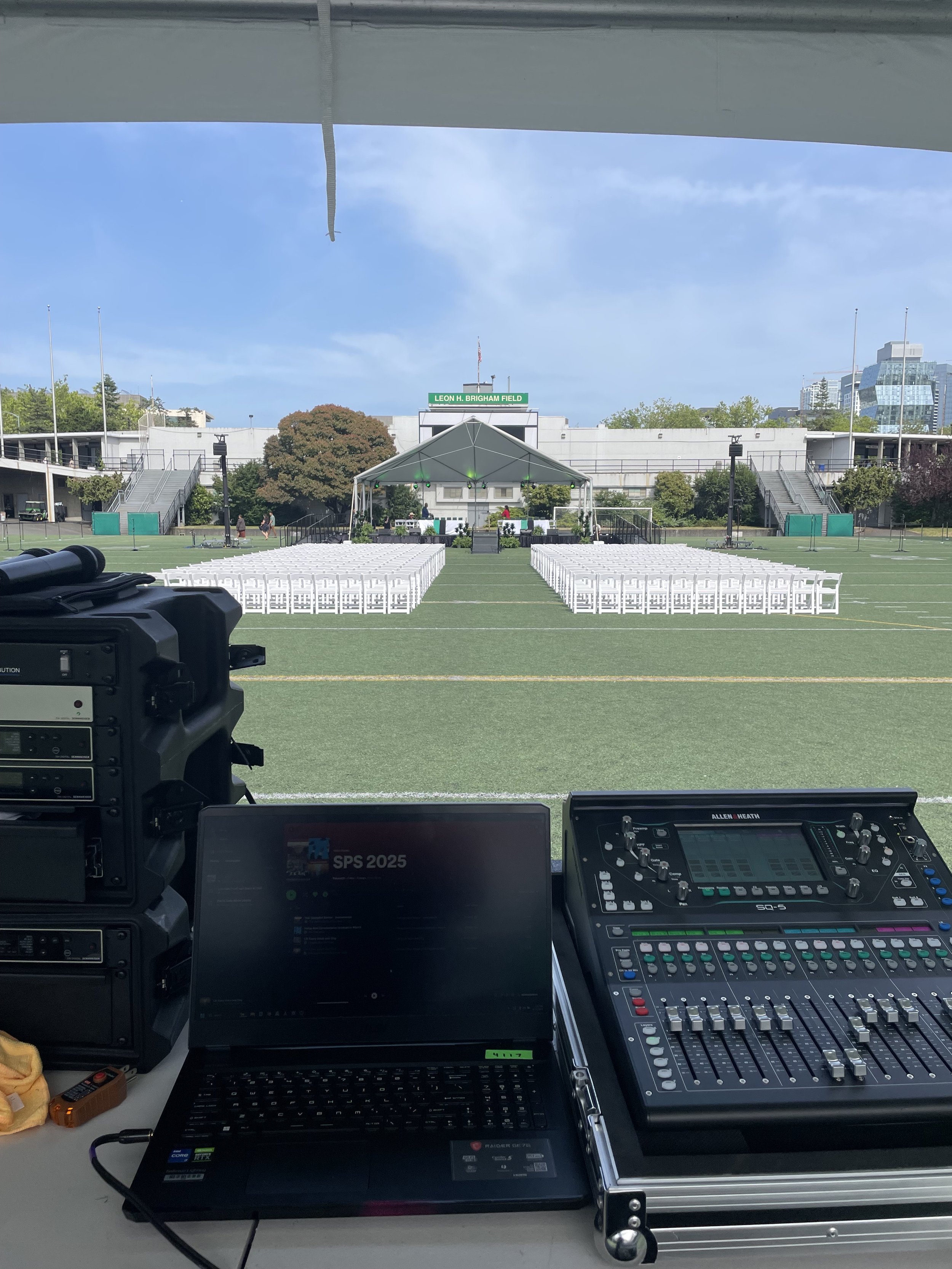 Sound and lighting control equipment on a table in a studio view of a field with chairs and a stage in the background.