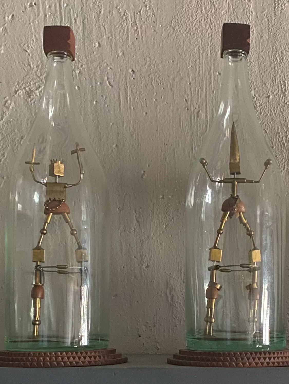 Two glass bottles with metal and wood sculptures inside, resembling humanoid figures with outstretched arms, placed on wooden bases against a textured wall.