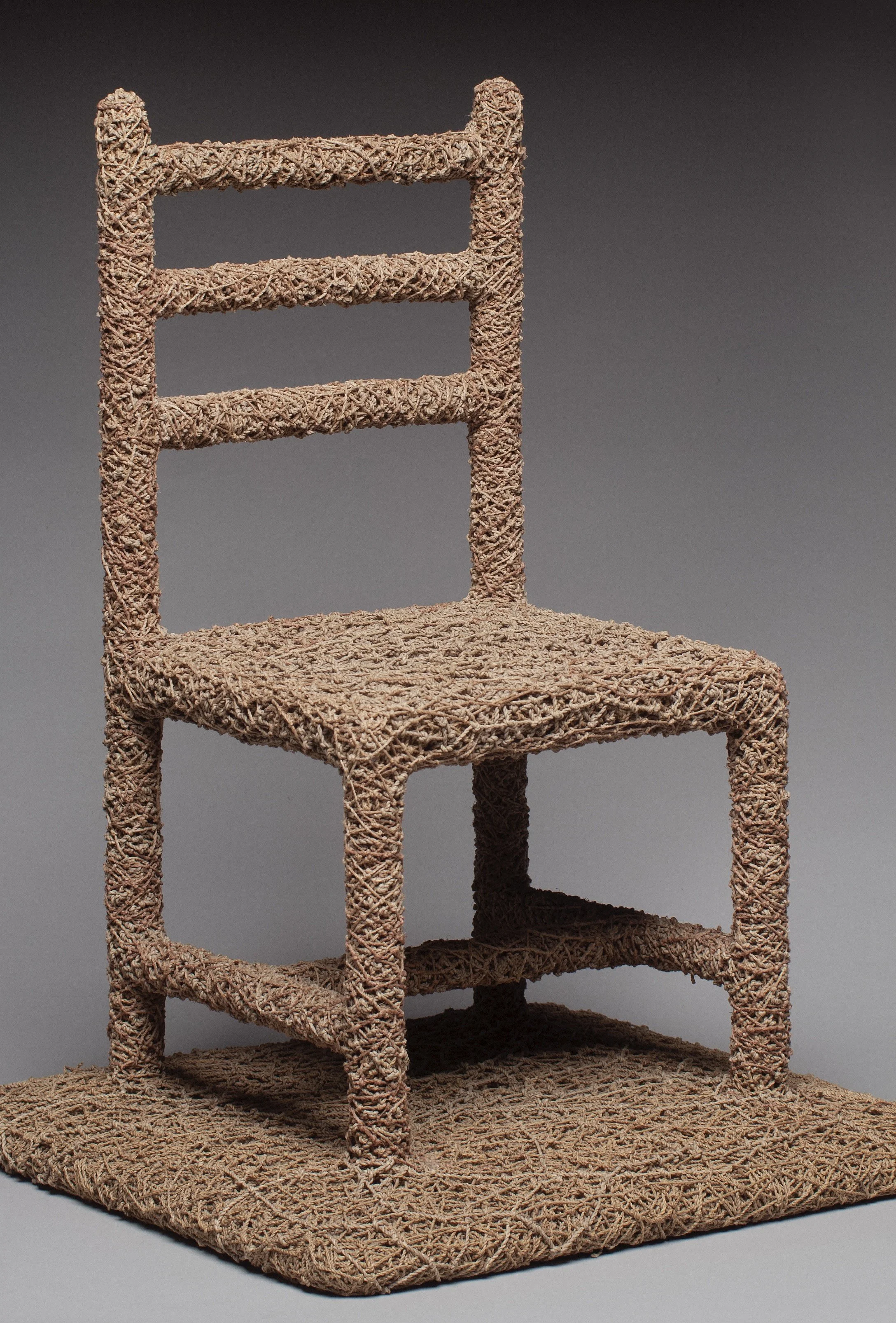 A chair made of intertwined natural materials, resembling straw or rattan, with a large square base and a smaller square seat, and a backrest with three horizontal slats.