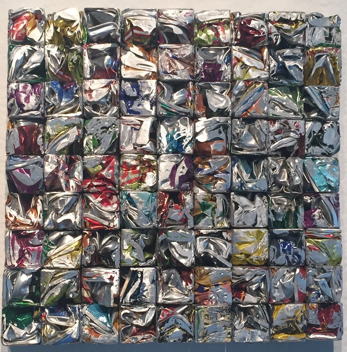 Artwork composed of crushed, metallic soda cans arranged in a grid pattern.