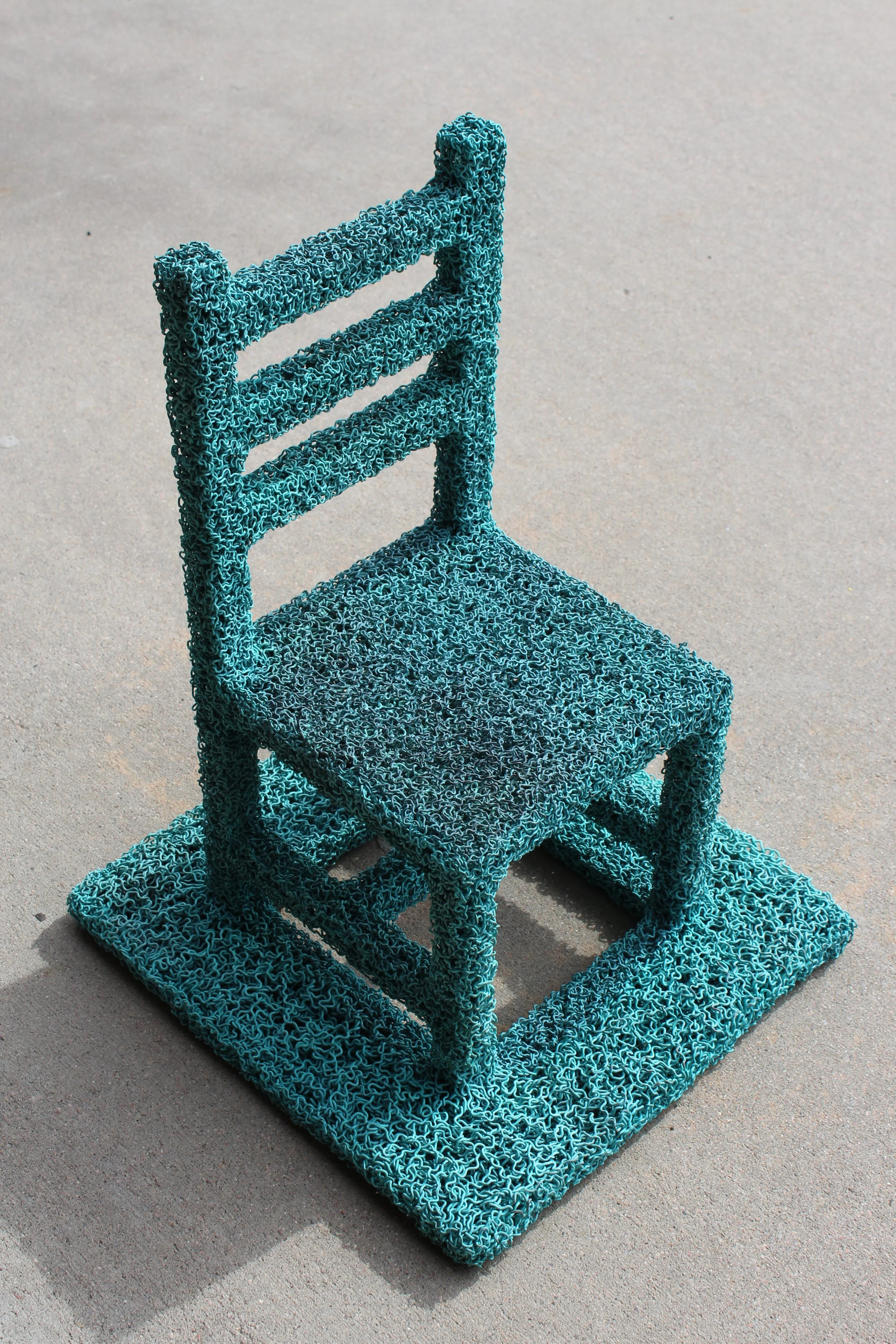 A chair made from blue metal shavings, designed with three horizontal bars on the back, a solid seat, and standing on a textured, rectangular base. The chair is on a concrete surface with shadows cast by sunlight.