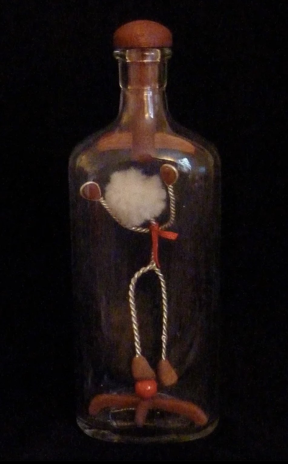 A decorative glass bottle with a wire frame of a person inside, featuring a cotton ball for the head, small orange circles for the ears, a red bow tie, and wooden beads for the shoes, set against a black background.