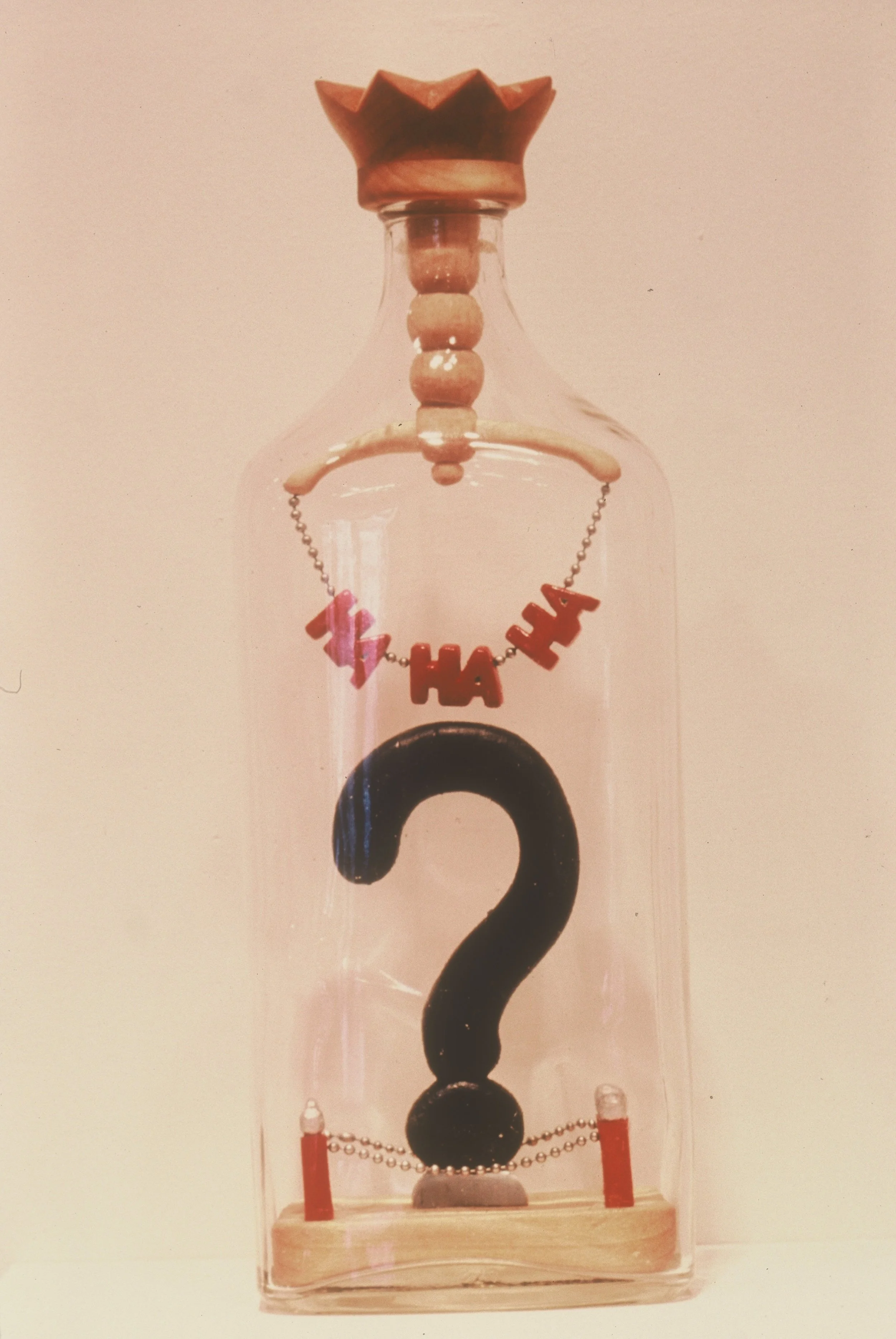 Glass bottle with a wooden lid, decorative beads on top, and a large black question mark inside. The bottle has a string of beads with the word "HAHA" and star-shaped beads hanging. The background is plain and light-colored.