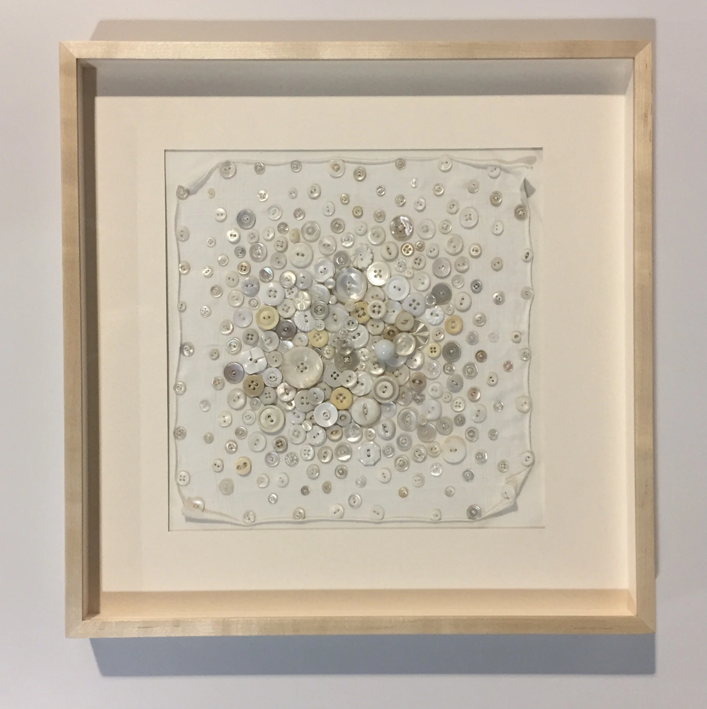 Framed artwork of various white, silver, and beige buttons arranged on a fabric background.