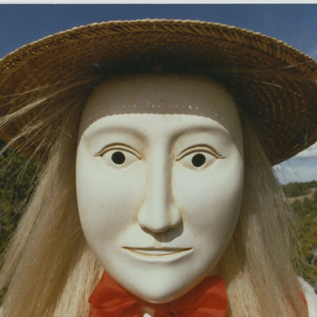A close-up of a white mask with realistic human facial features, wearing a wide-brimmed straw hat and an orange garment with a bow, outdoors with trees and sky in the background.