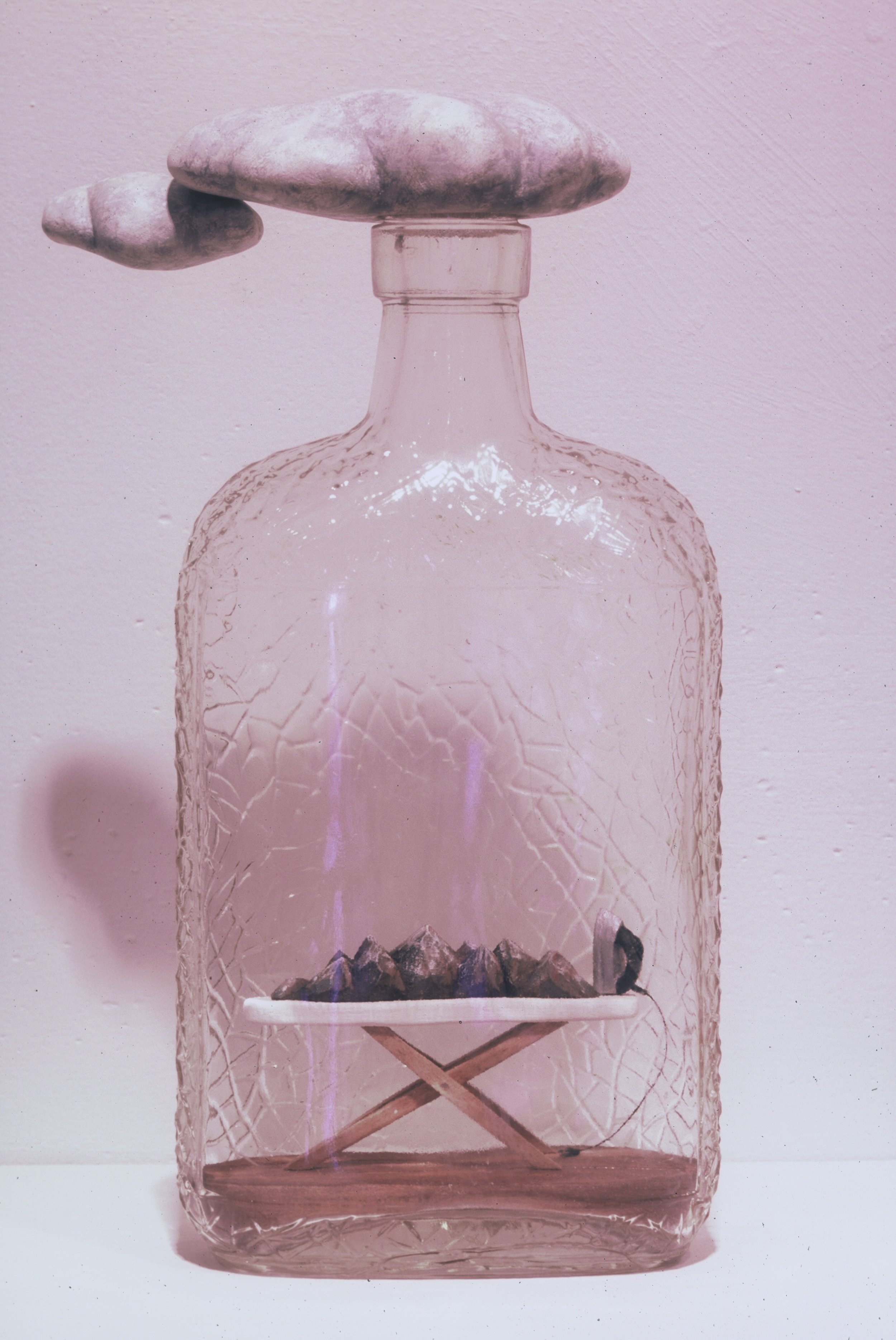 A transparent pink glass bottle with a textured surface, topped with a large rock, a smaller rock and a cloud-shaped rock on top. Inside, there is a small shelf holding black stones, and a tiny black bird perched on the edge.
