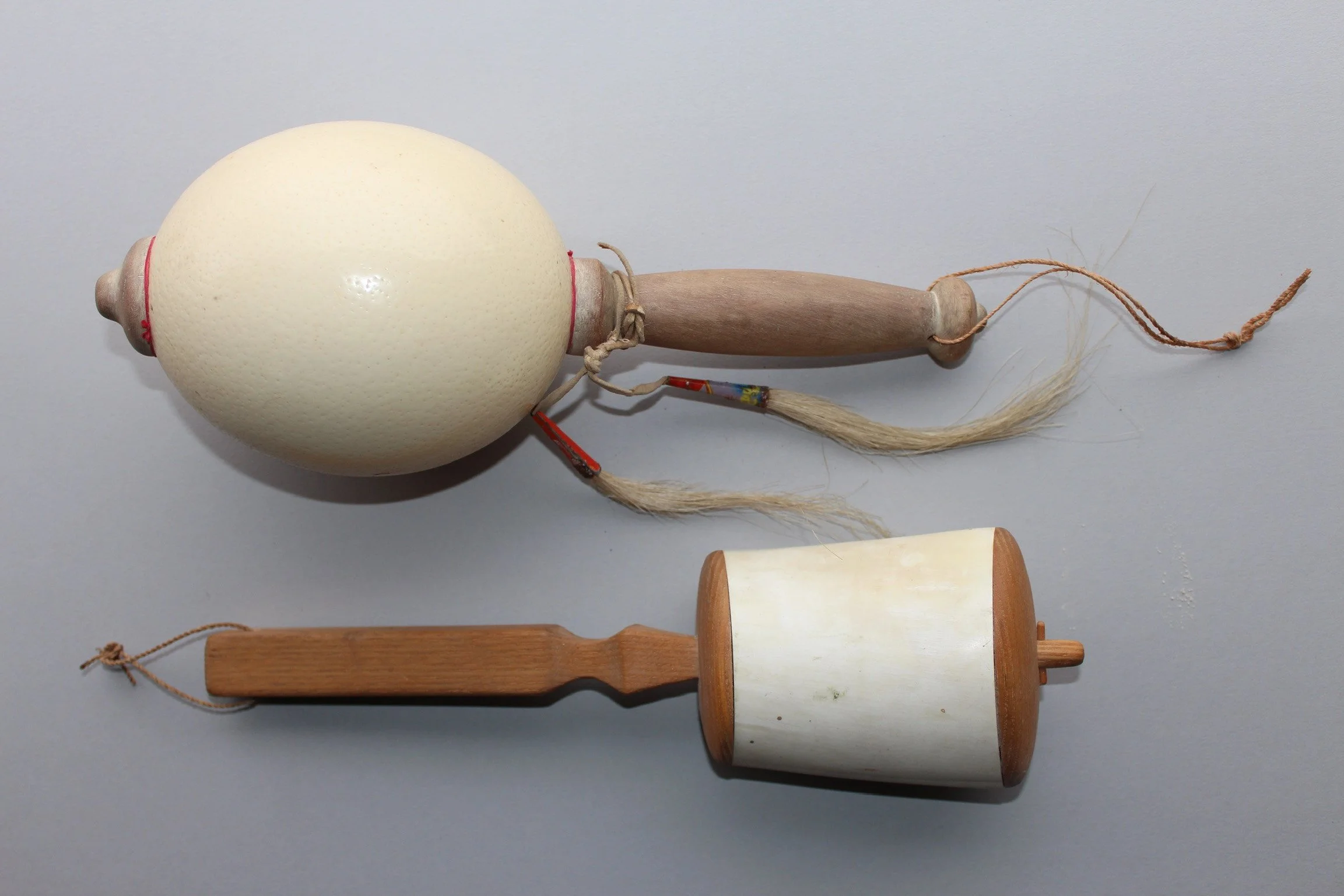Two traditional musical instruments, a wood and ivory rattle with a large round top, and a small wood and ivory drum, both on a plain gray background.