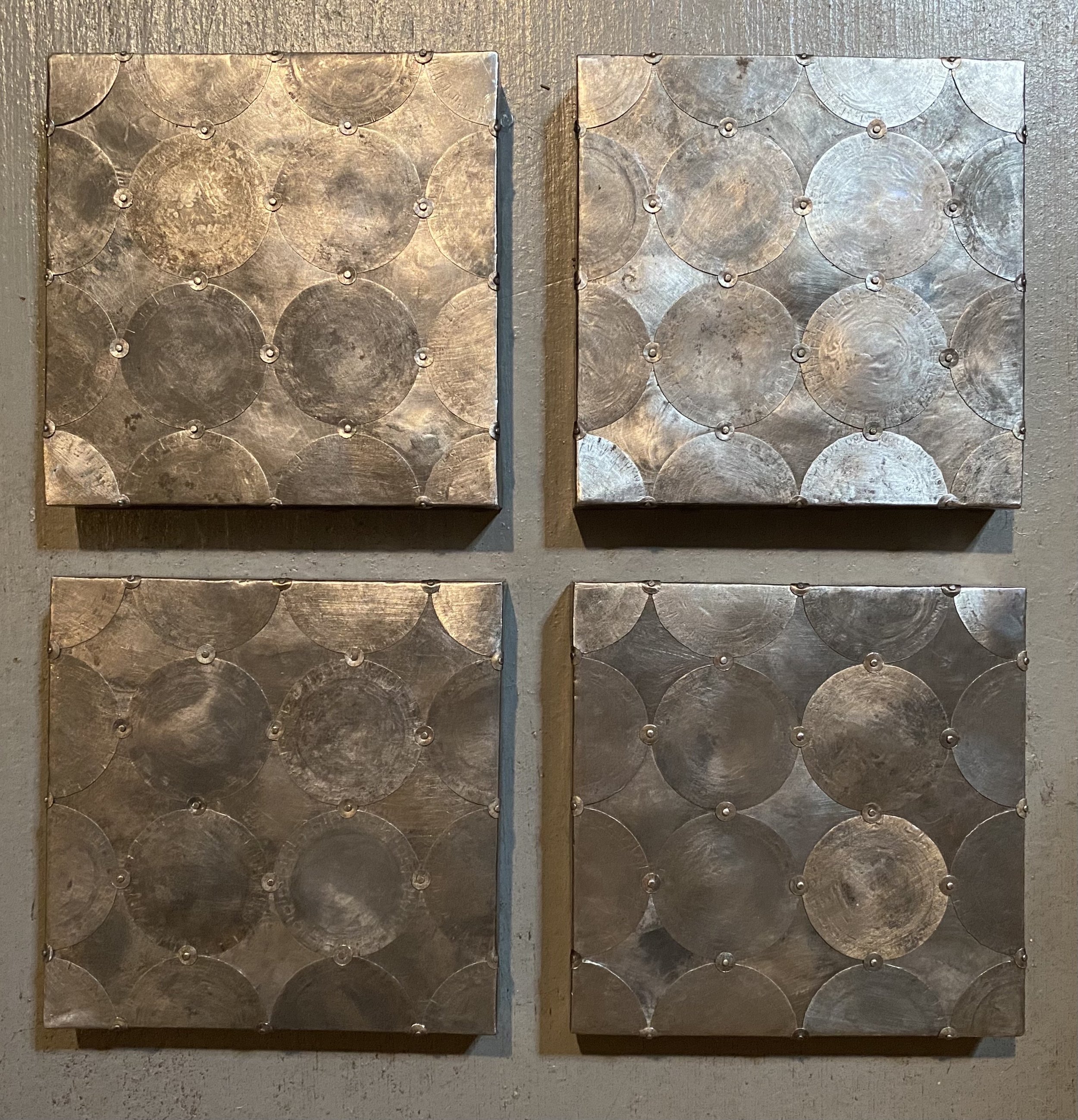 Four square metal wall panels with circular patterns, arranged in a 2x2 grid, mounted on a wall.