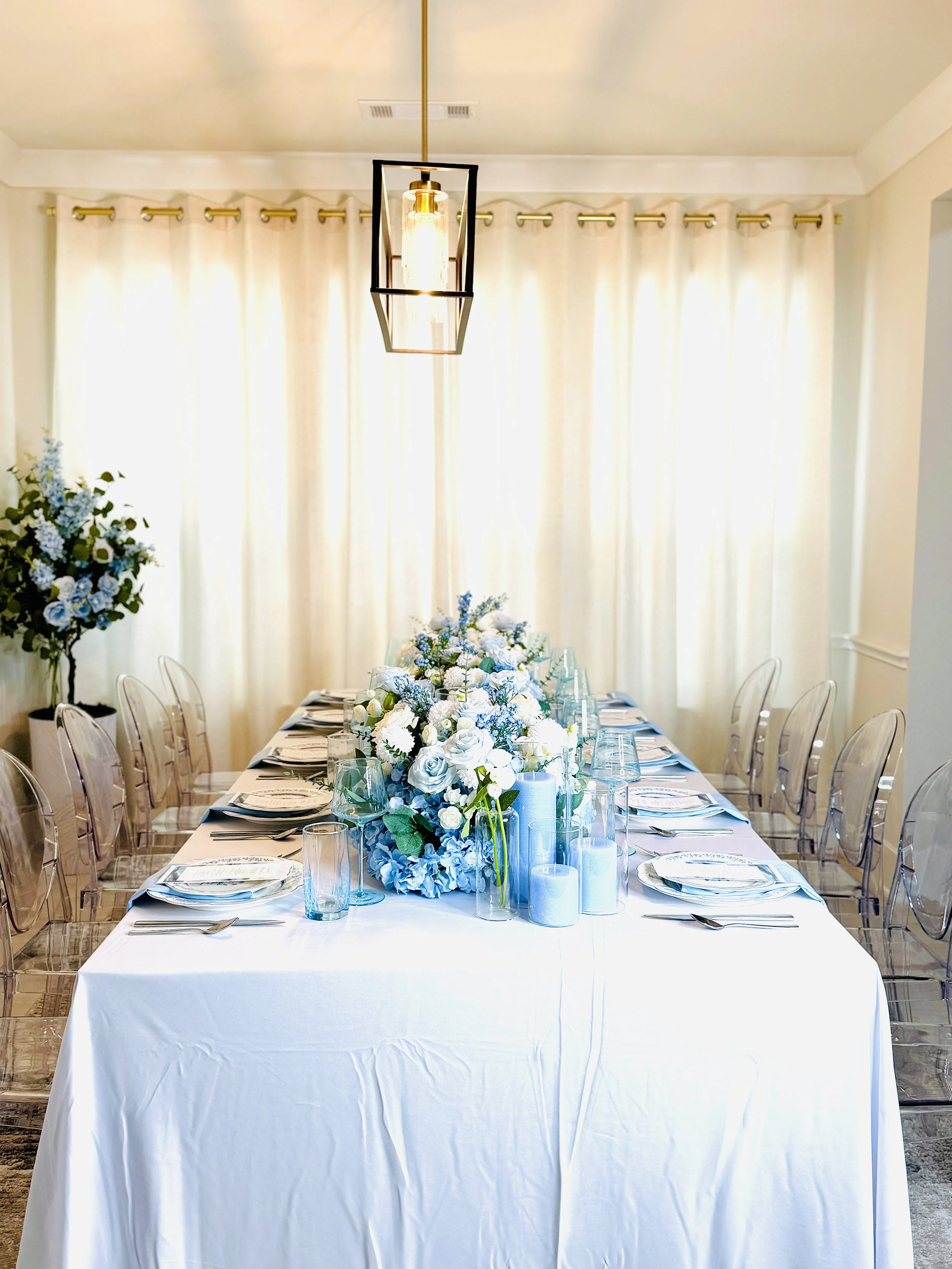 Elegant dining table set for a formal event with white tablecloth, floral centerpiece in shades of white and blue, candles, and place settings, in a bright room with sheer white curtains.