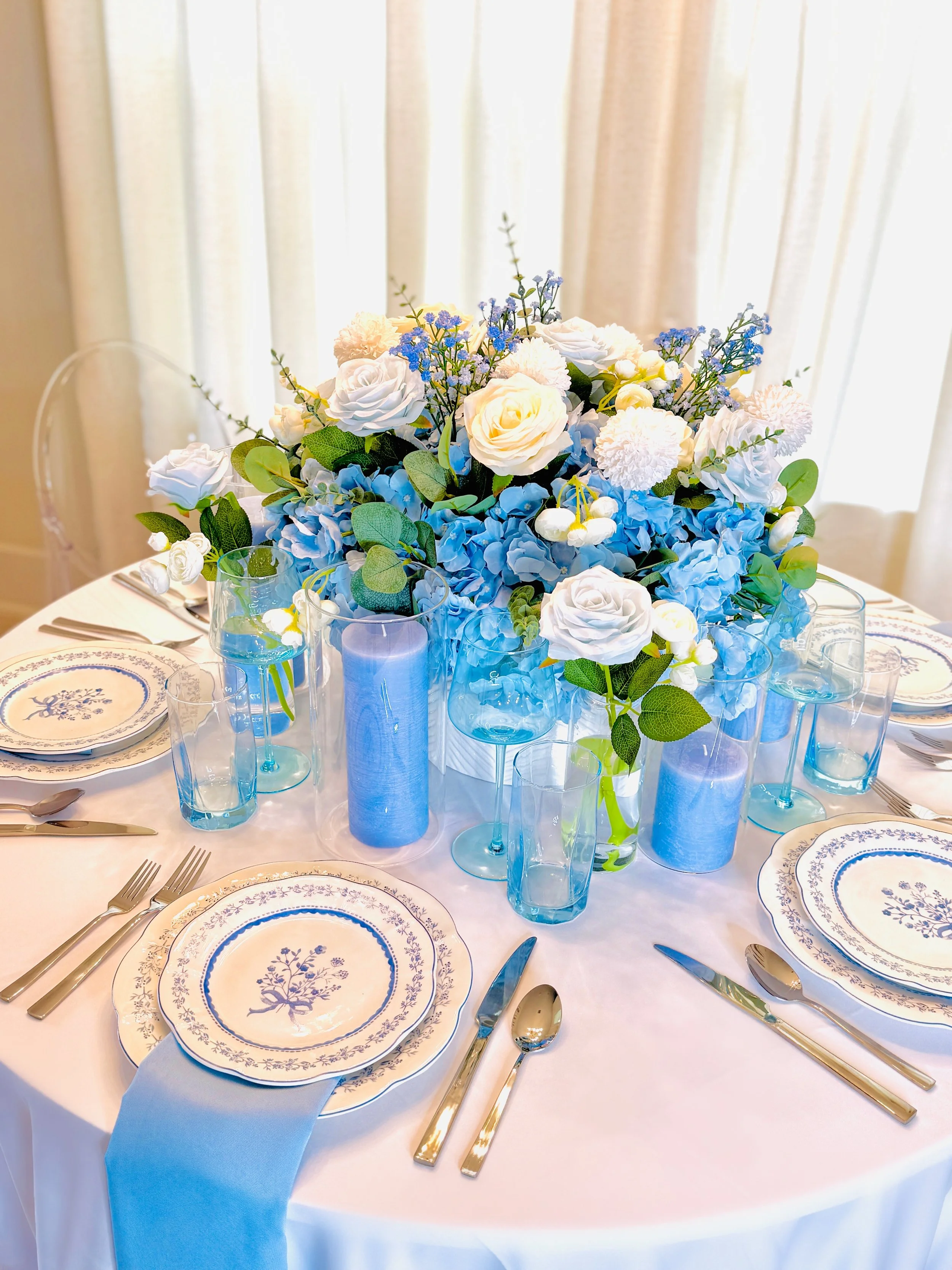 A table set for a formal event with blue and white floral centerpiece, blue candles, and matching china plates and glassware.