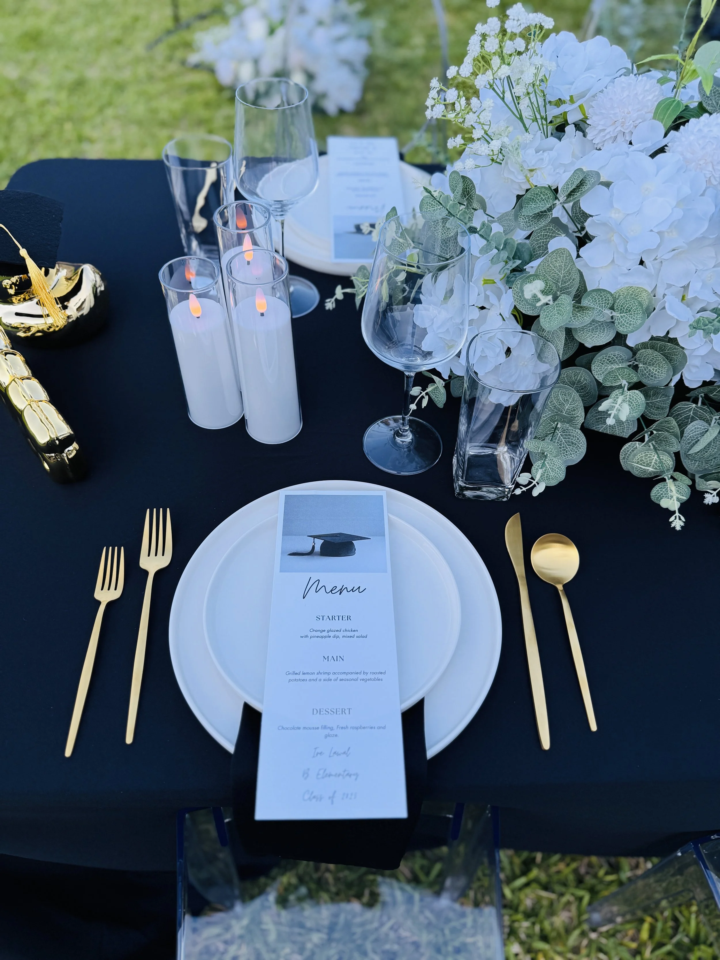 Elegant outdoor dining table with black tablecloth, gold cutlery, white plates, a menu with a graduation cap image, white floral centerpiece, clear glassware, and white candles.