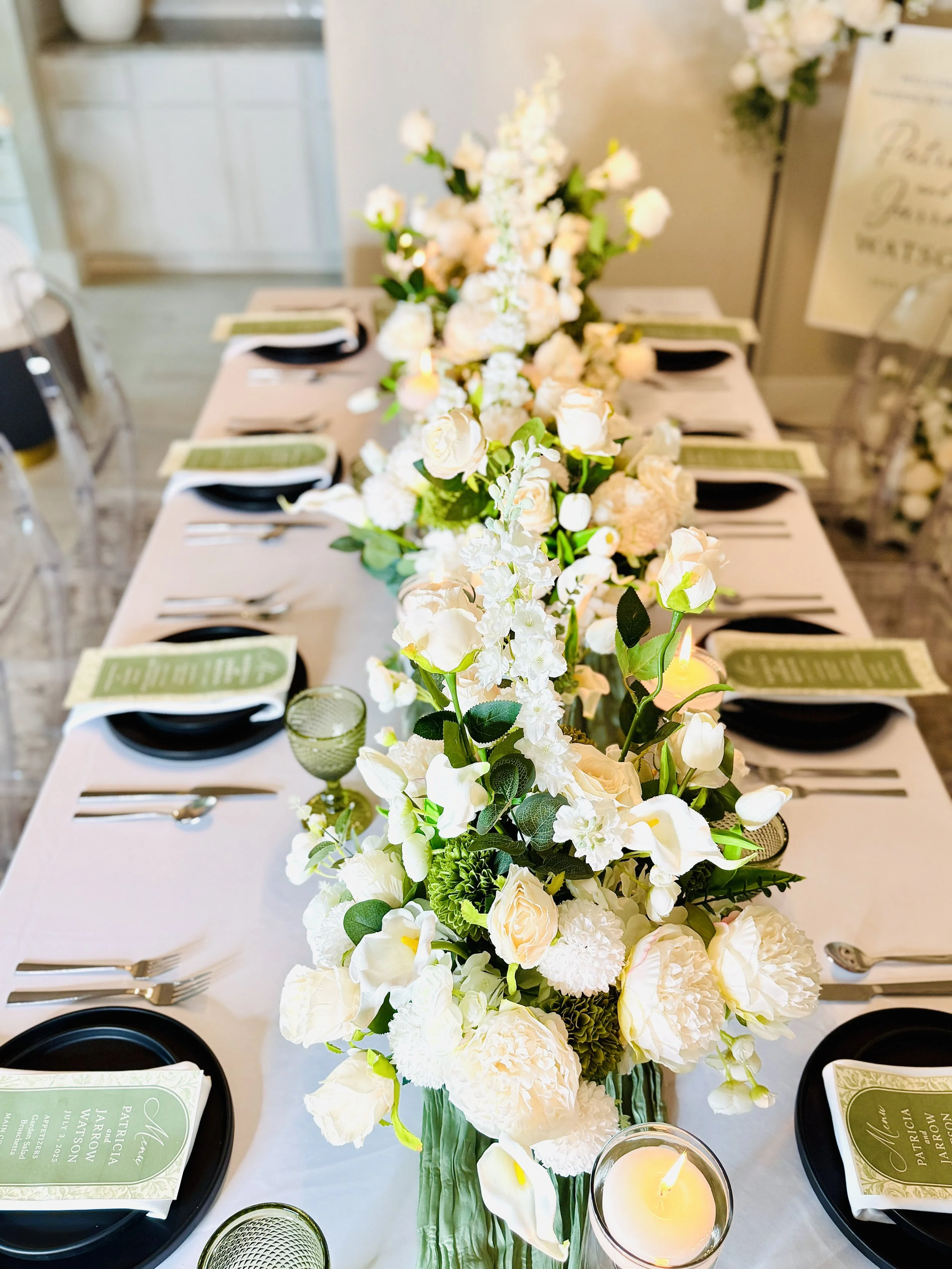 Elegant table setting for a wedding with a long floral centerpiece of white and cream roses, tulips, and other white flowers, with candles and place settings including black plates and green menus.