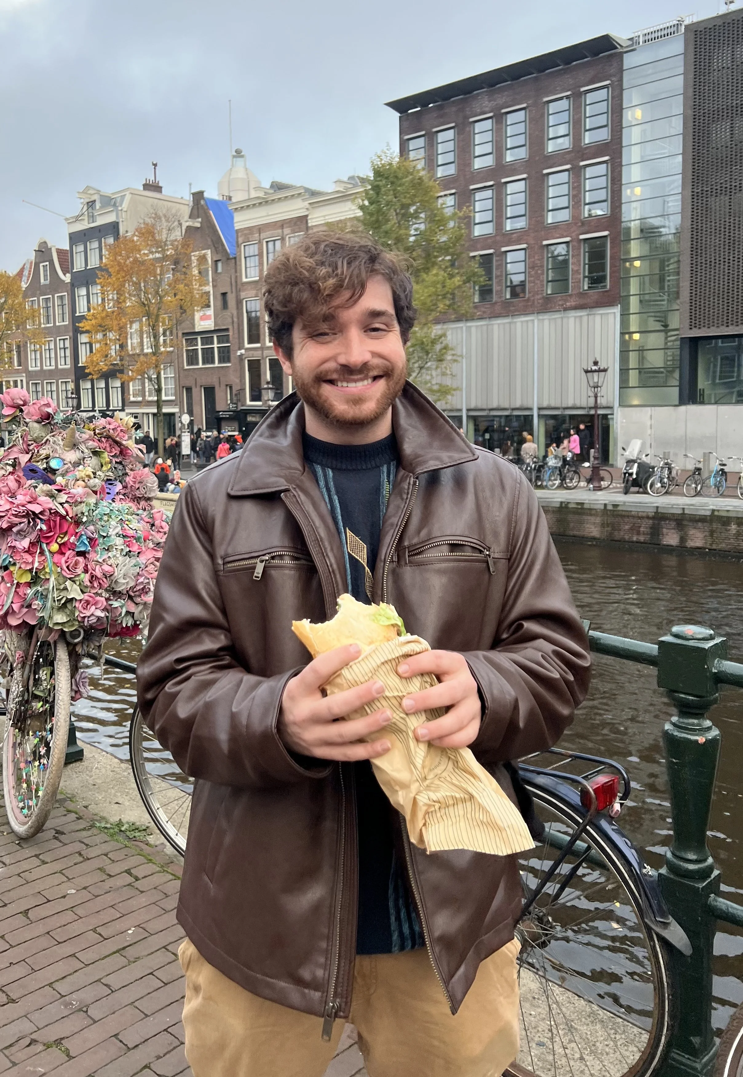 A smiling man holding a sandwich wrapped in paper on a riverside sidewalk in an urban area with bicycles parked nearby and colorful buildings in the background.
