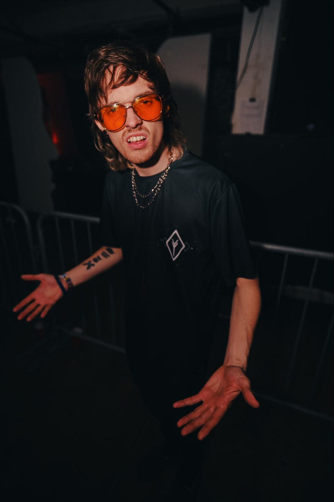 A man with long hair and sunglasses wearing a black shirt with a logo, standing behind a metal barrier at a dark indoor event, making a confused or annoyed facial expression.