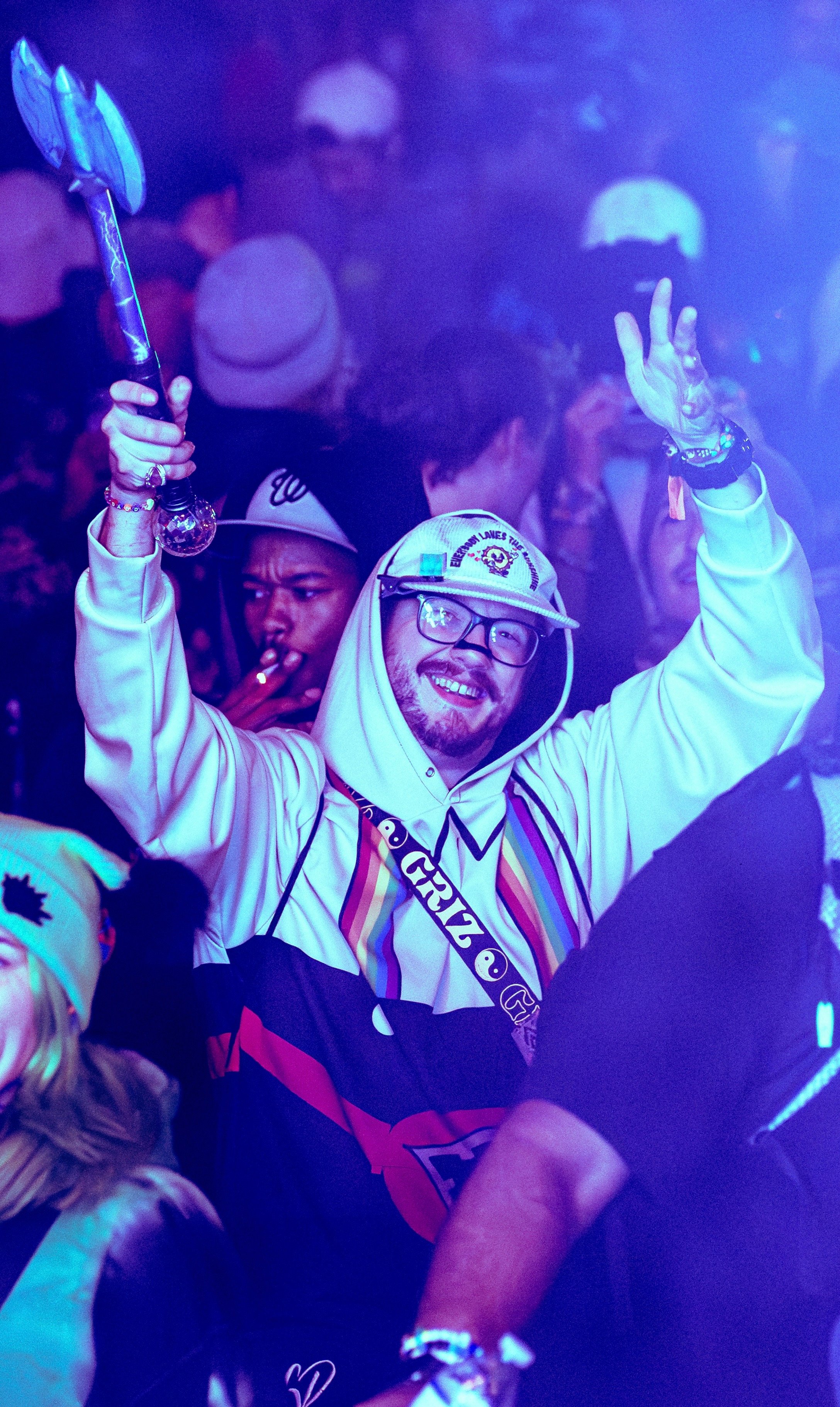 A group of people dancing and celebrating at a party with purple lighting, some wearing hats and accessories, with one person in the foreground smiling and holding a prop, wearing a hoodie and glasses.