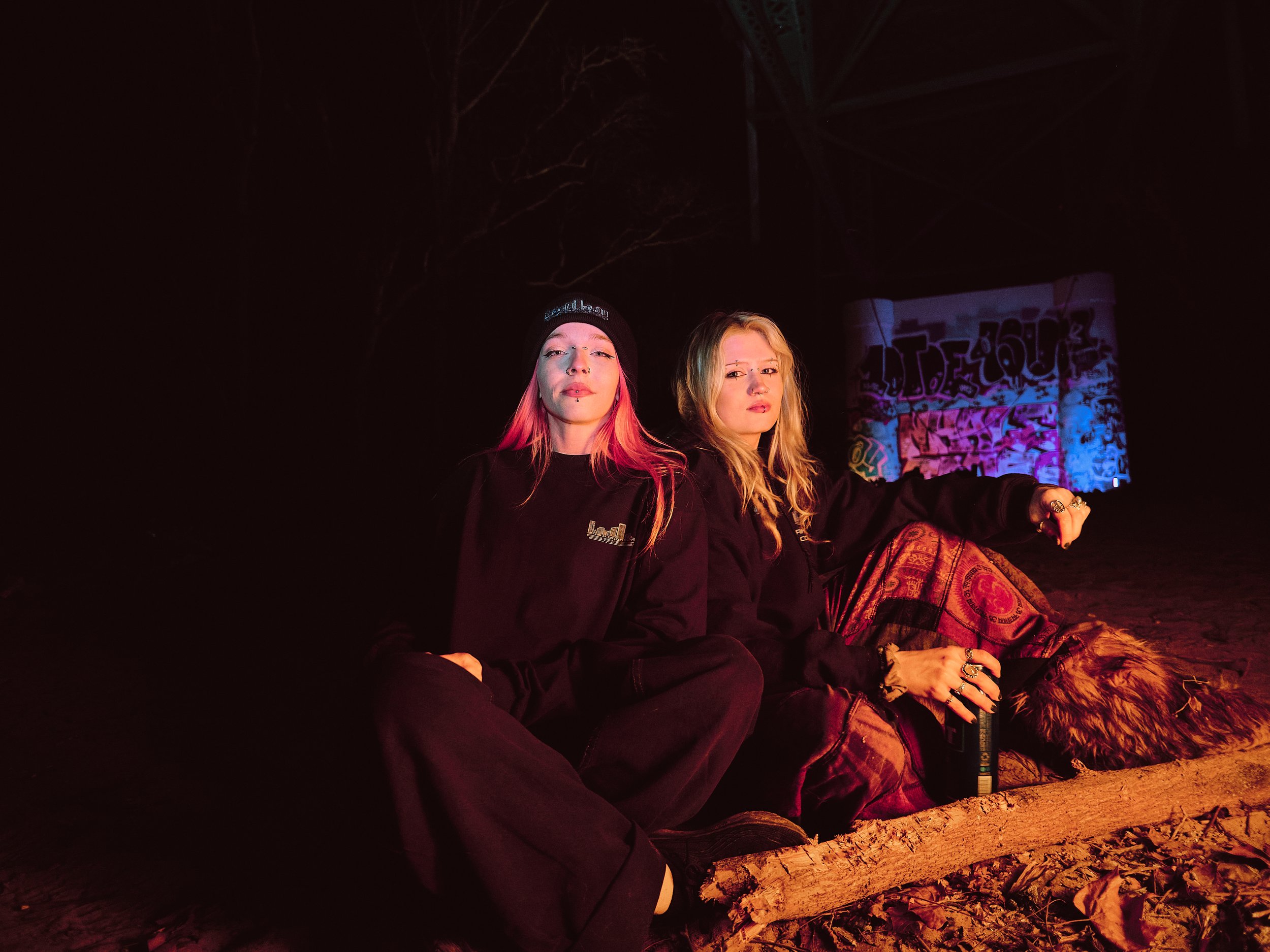 Two young women sitting on the ground at night, illuminated by colorful lighting, with graffiti in the background.