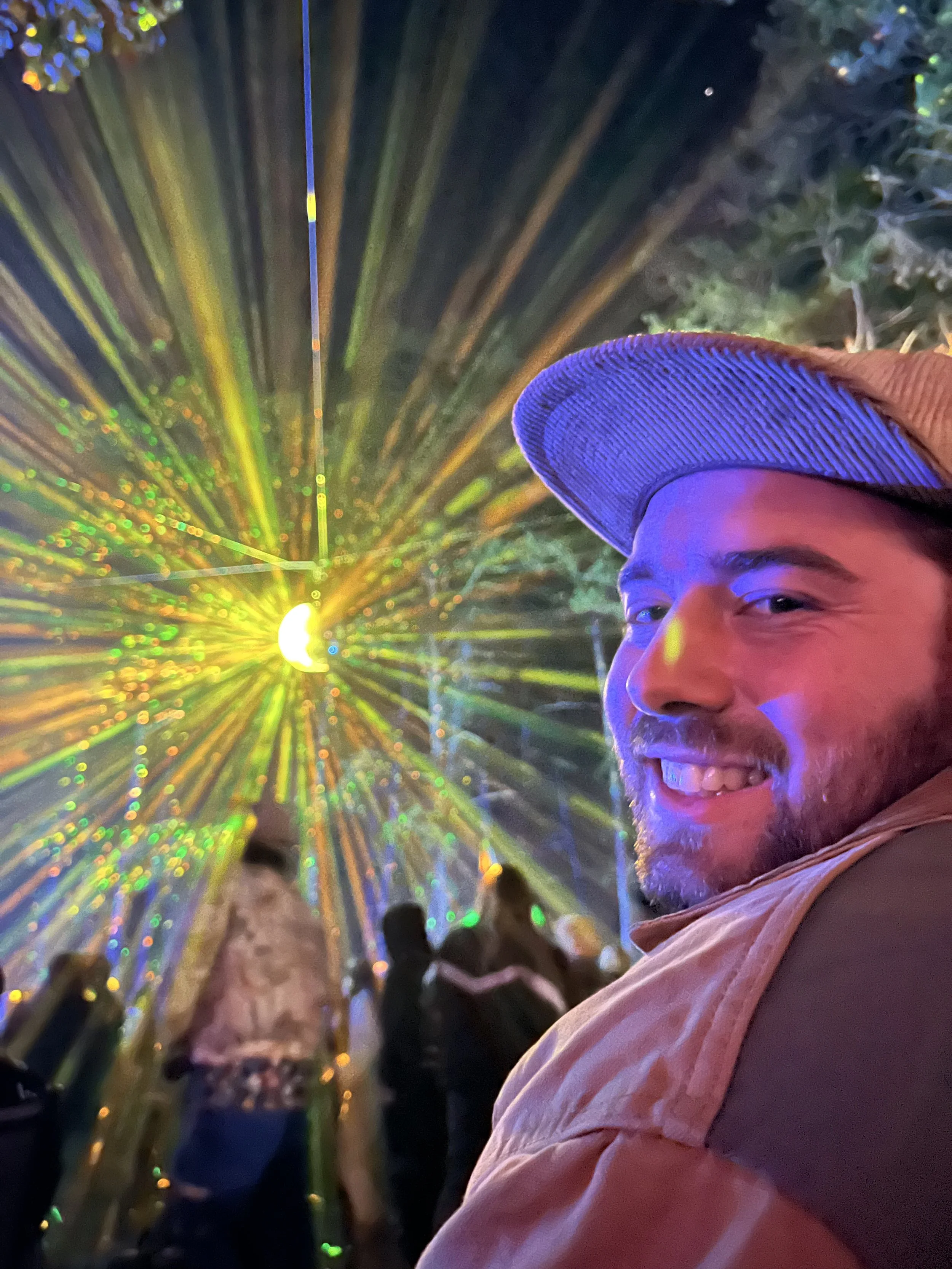 A man smiling at a colorful laser light display while wearing a baseball cap and outdoor jacket, with blurred people in the background.