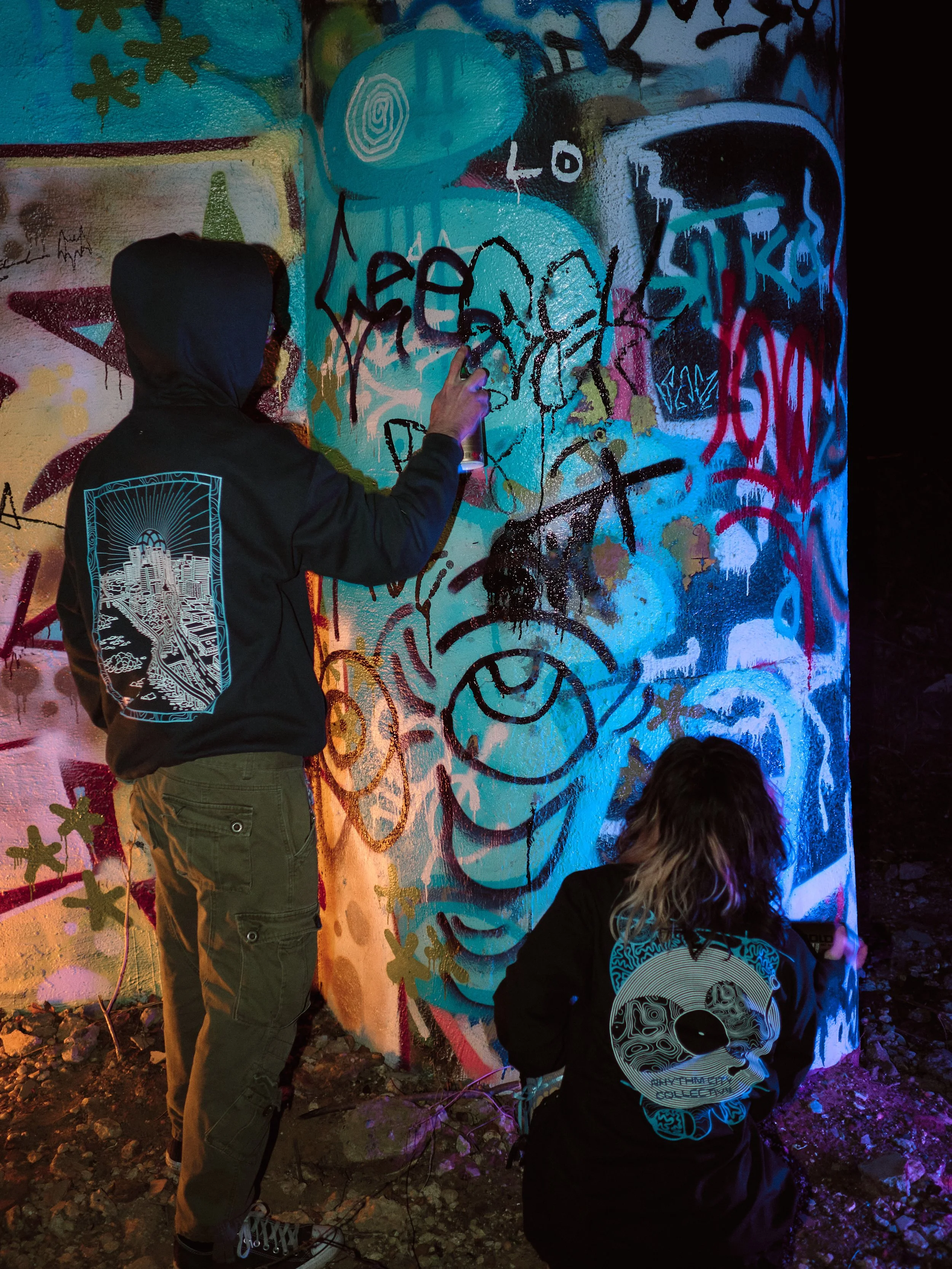 Two people creating graffiti art on a wall at night. One person is standing and appears to be spray painting, while the other is crouched down. The wall is covered in colorful graffiti, including various tags, symbols, and abstract designs.