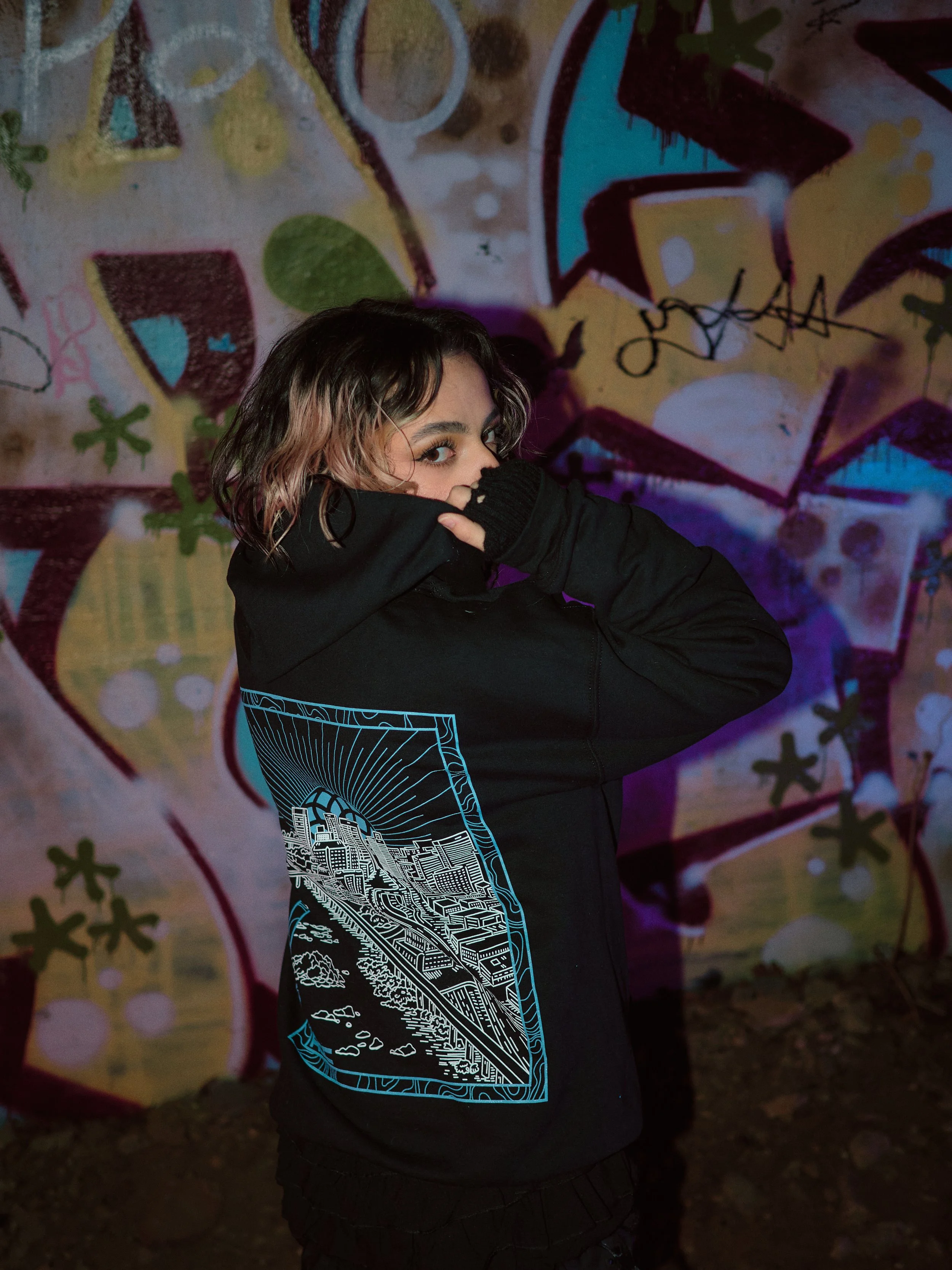 A woman with short, wavy hair partially covering her face is wearing a black hoodie with a graphic design of a cityscape on the back. She is standing in front of a colorful graffiti wall and holding the collar of her hoodie up to cover her nose.